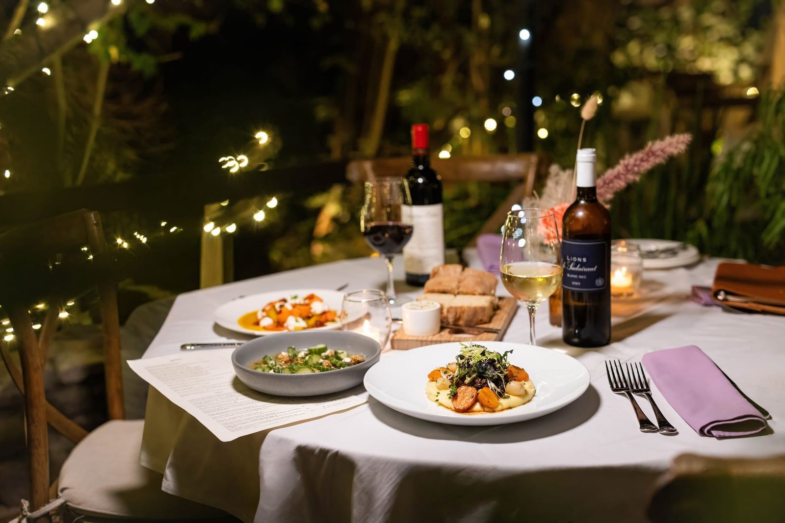 A round table set with plates of food and a bottle of wine in a garden lit with string lights at night.