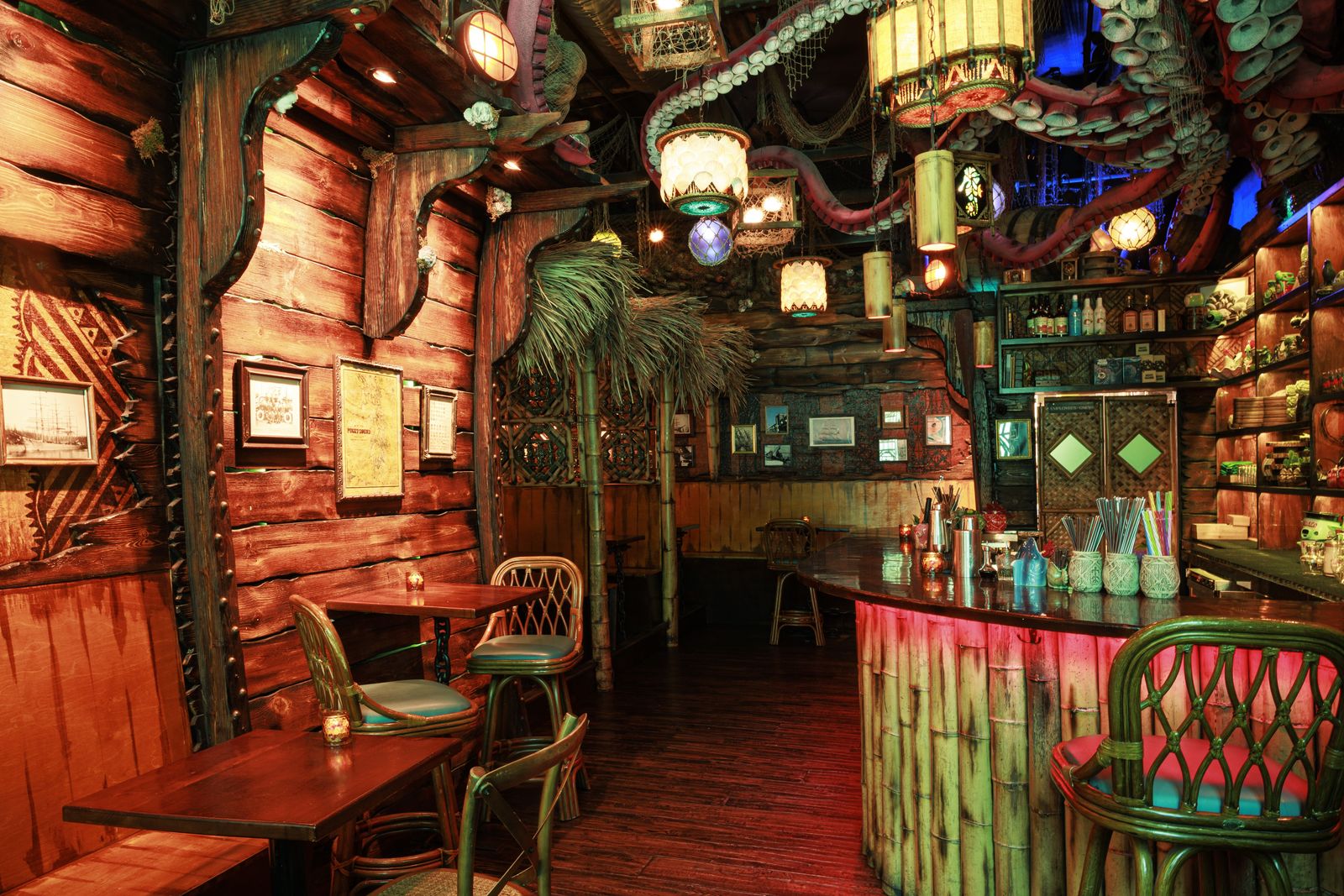 A tiki bar interior with carved wood walls, rattan chairs, a bamboo-fronted bar and hanging lamps.