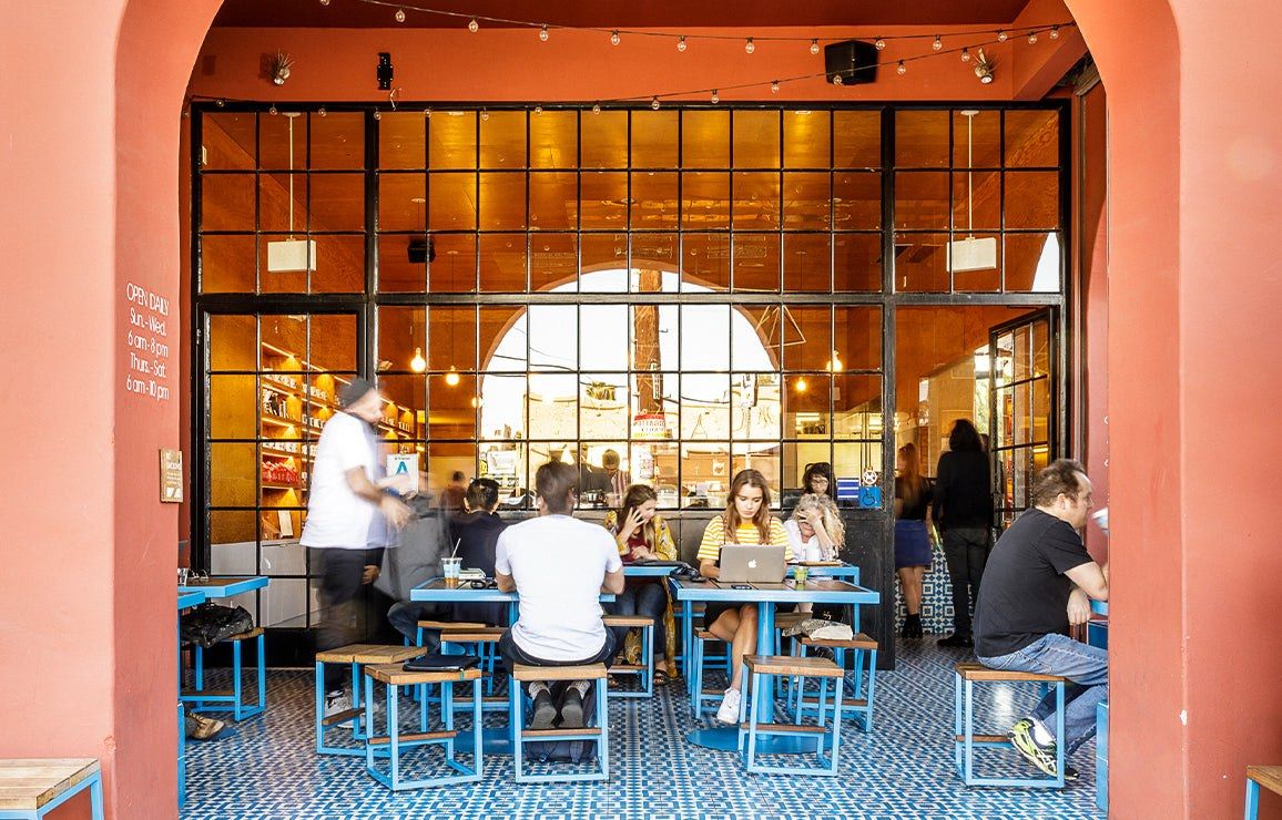 A cafe with a tall arched glass front and customers at tables with blue stools in an orange-walled room.