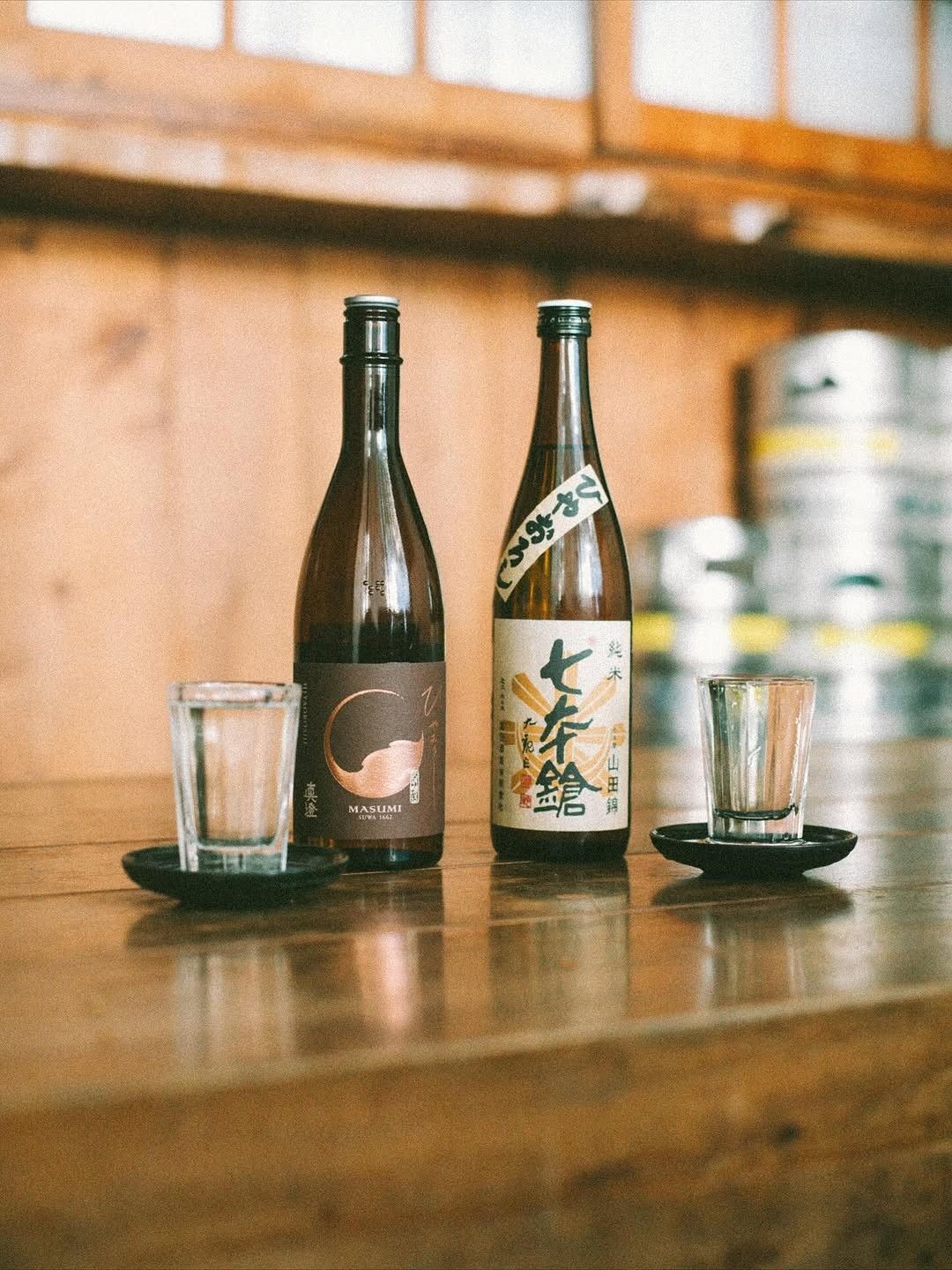 Two sake bottles and small cups on a wooden counter in front of a window.