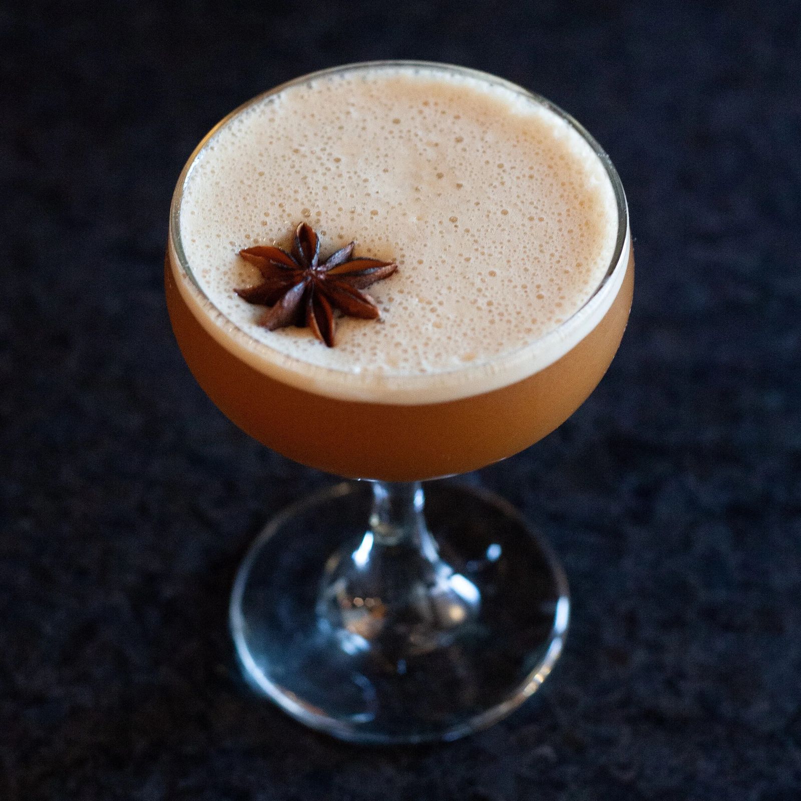 A frothy cocktail topped with a star anise pod in a coupe glass on a dark surface.