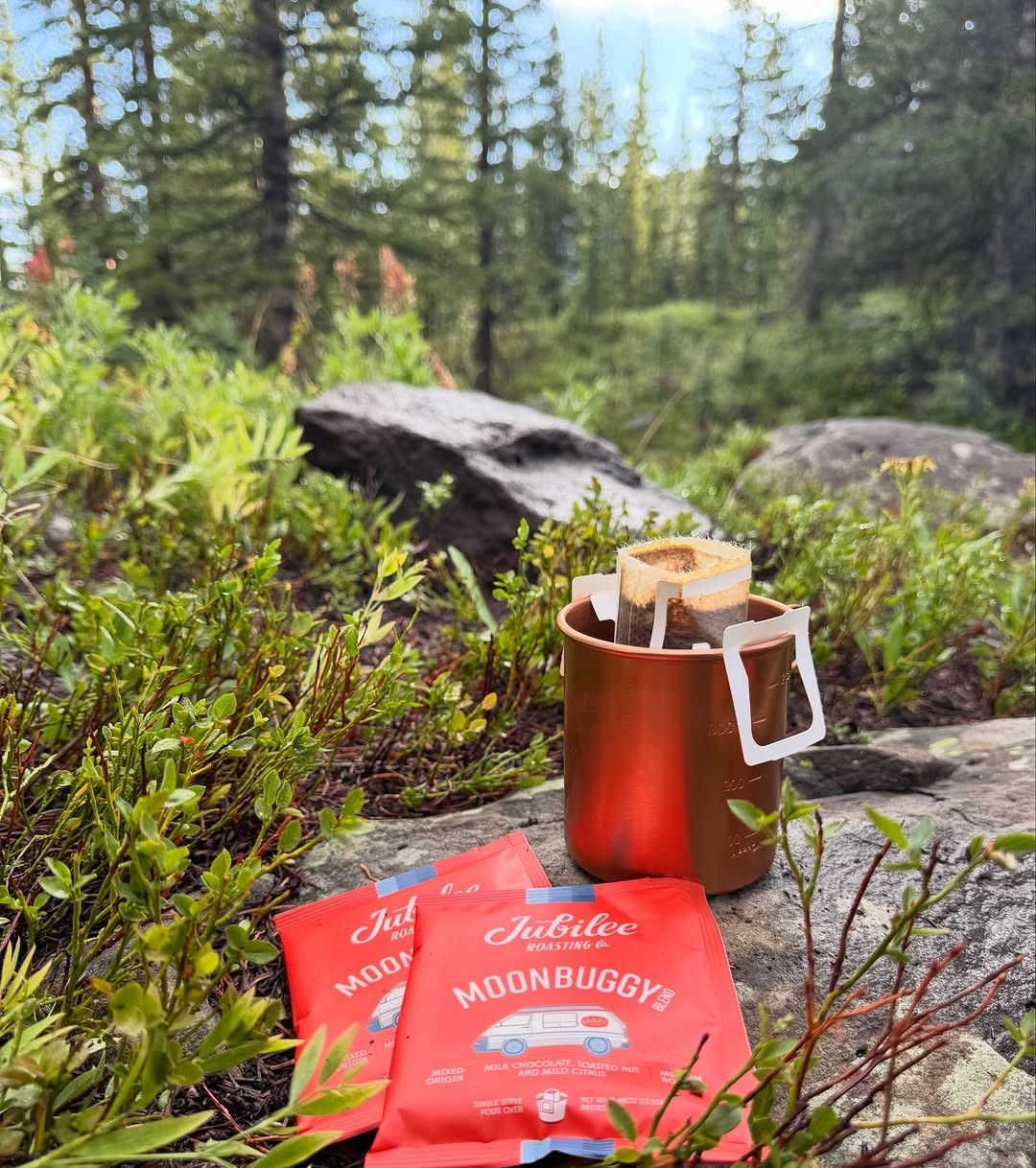 A red mug and packets of coffee on rocks and moss among pine trees on a trail.