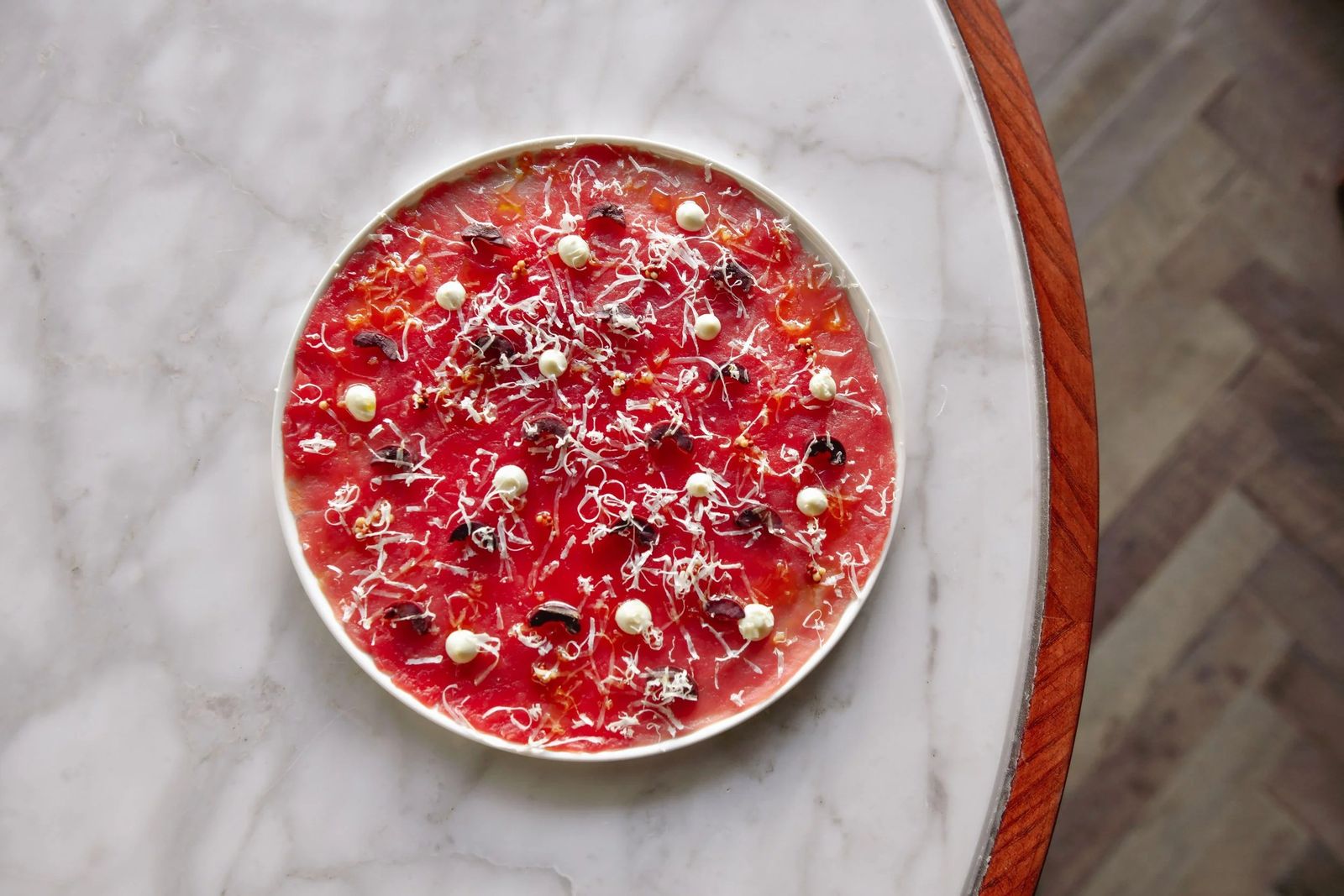 Overhead view of a round plate of red crudo topped with small white dots on a marble table