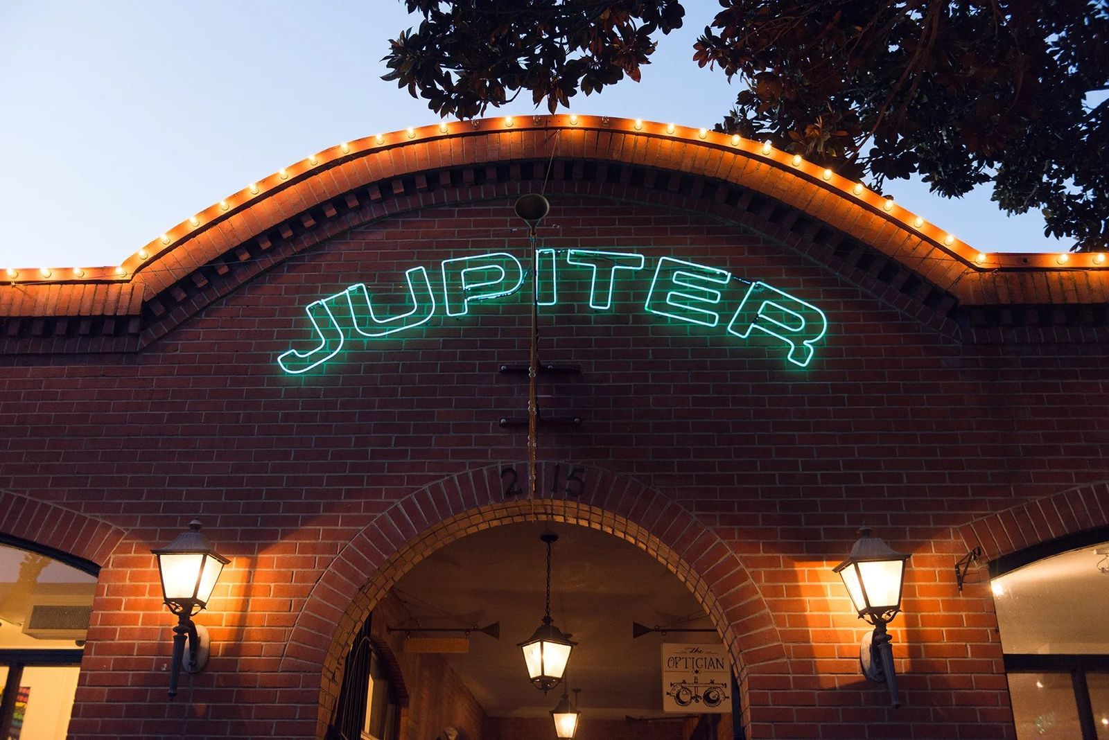 A brick archway entrance with a green neon Jupiter sign and warm lamps at night.