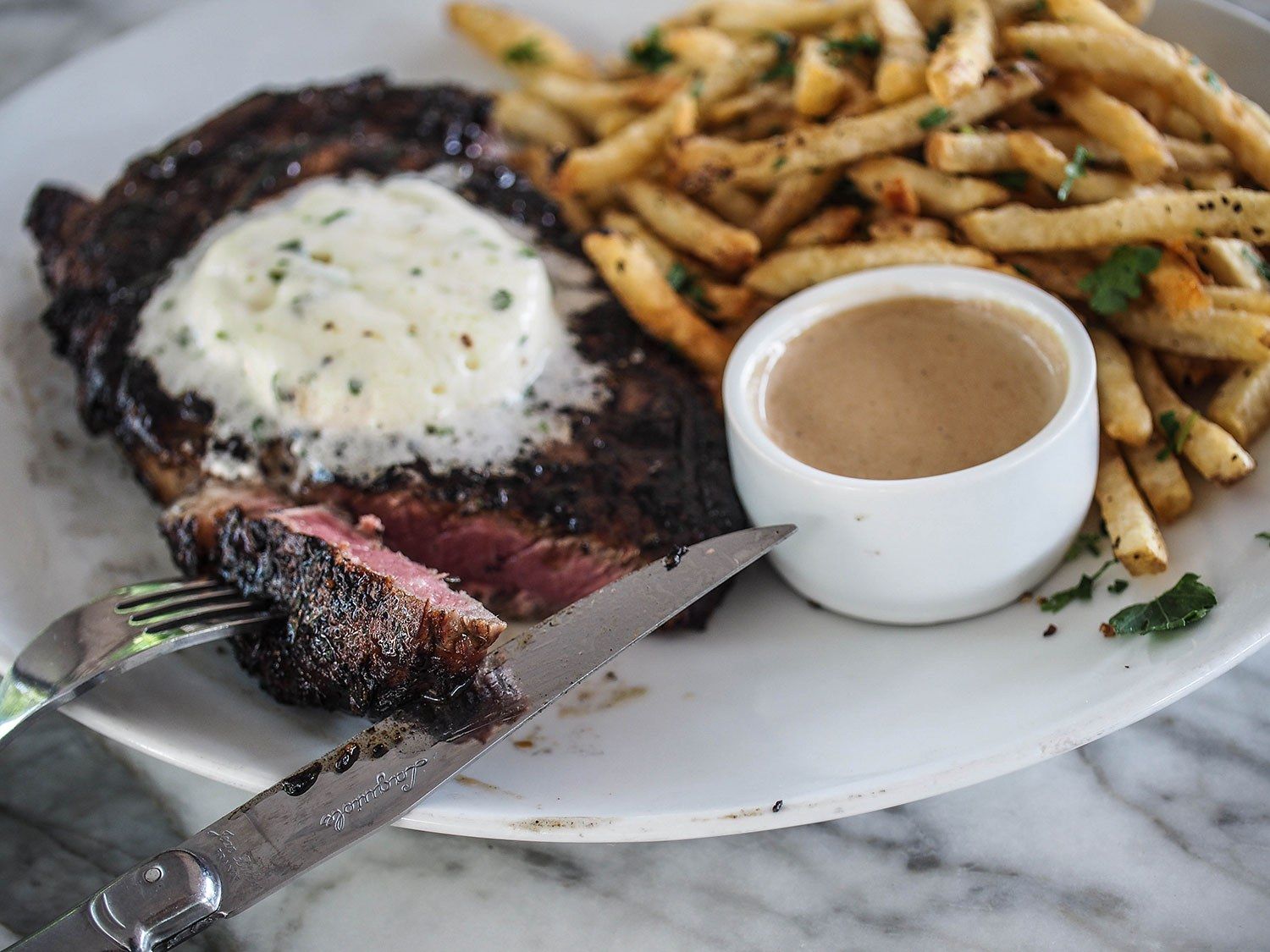 A steak with melted butter and a heap of fries beside a small dish of sauce on a marble table.