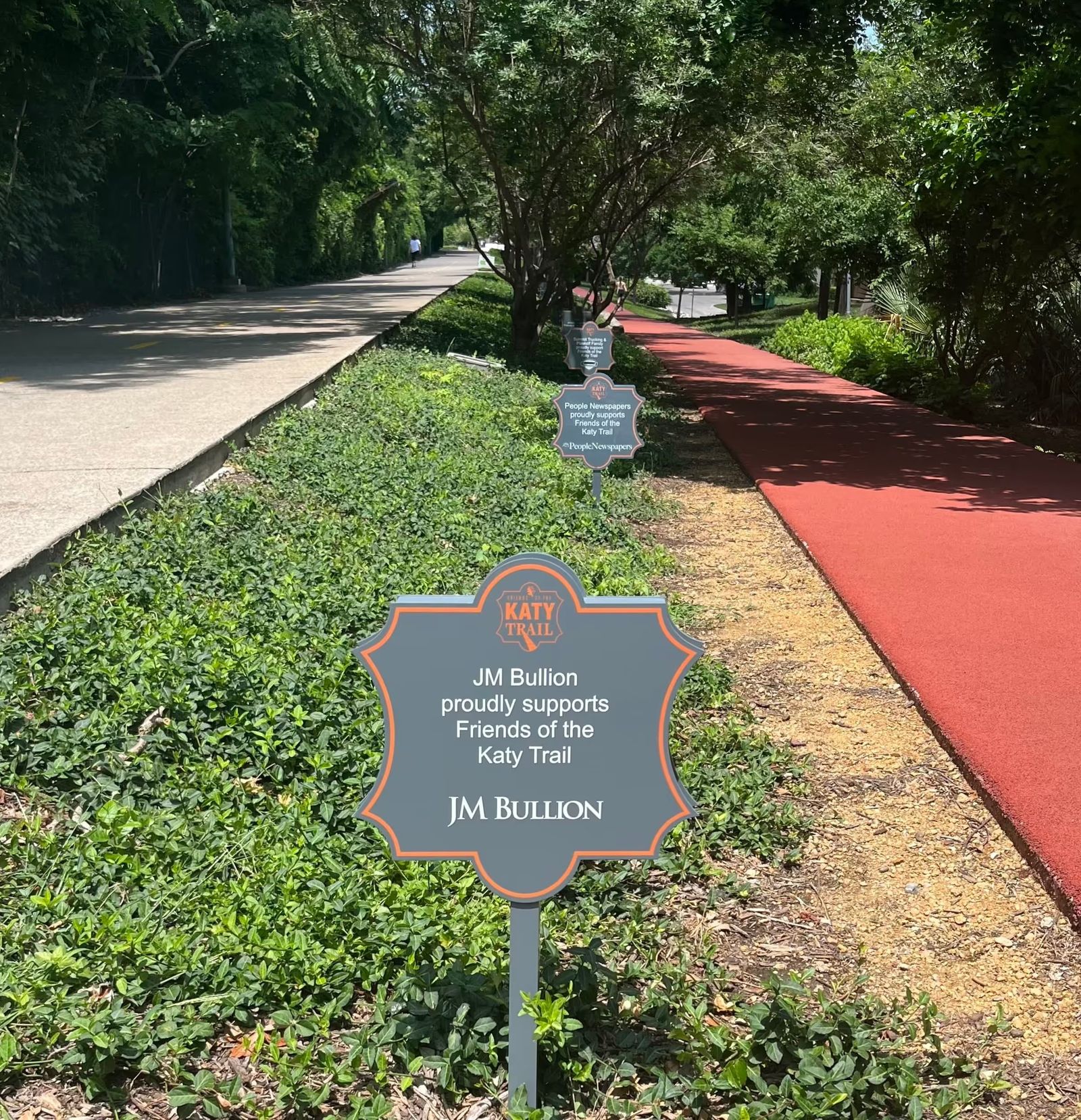 A paved walking path beside a red running lane, with plantings and a trail sponsor sign.