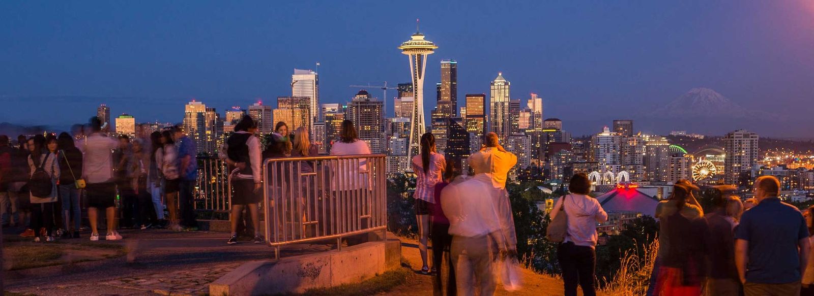 A crowd of visitors at the viewpoint railing at dusk, facing the Seattle skyline and the Space Needle.