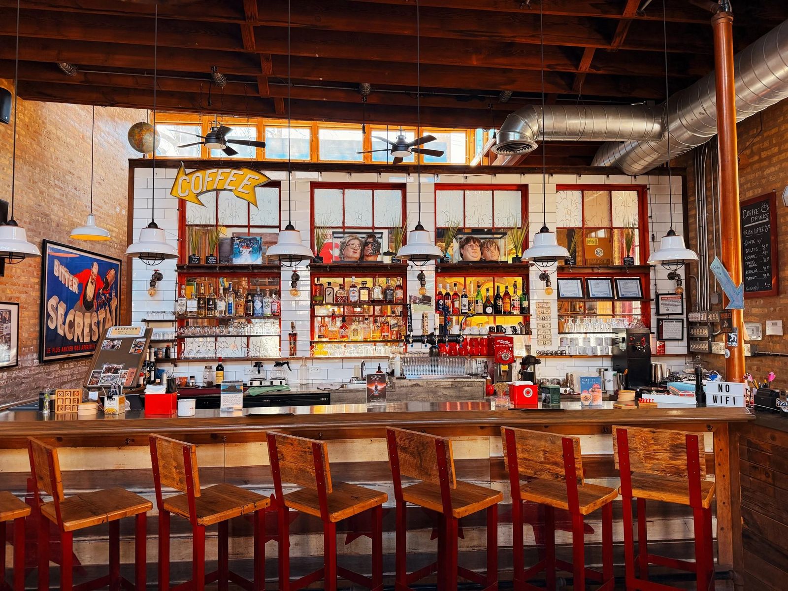A bar with high wooden stools and shelves of bottles under a ceiling of beams and ductwork in a brick room.
