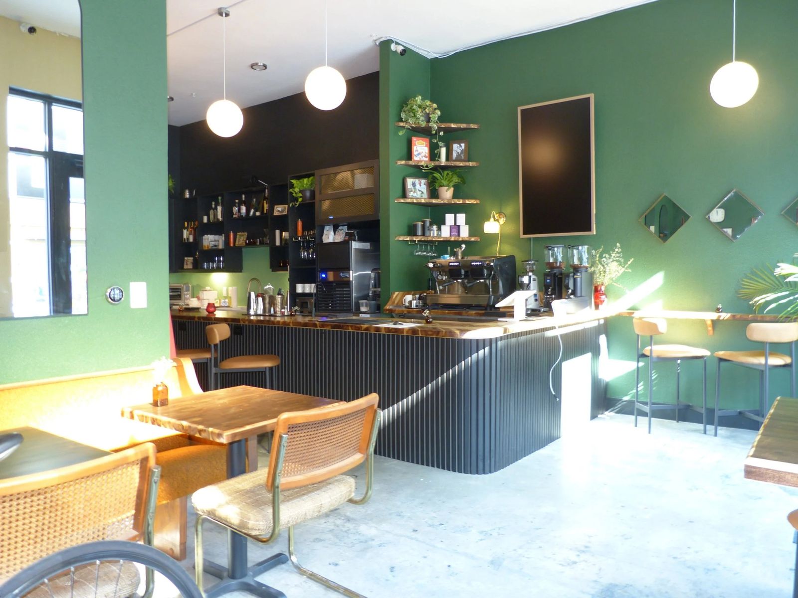 A cafe interior with green walls, a counter with espresso equipment and shelves, and wood and cane chairs at small tables.