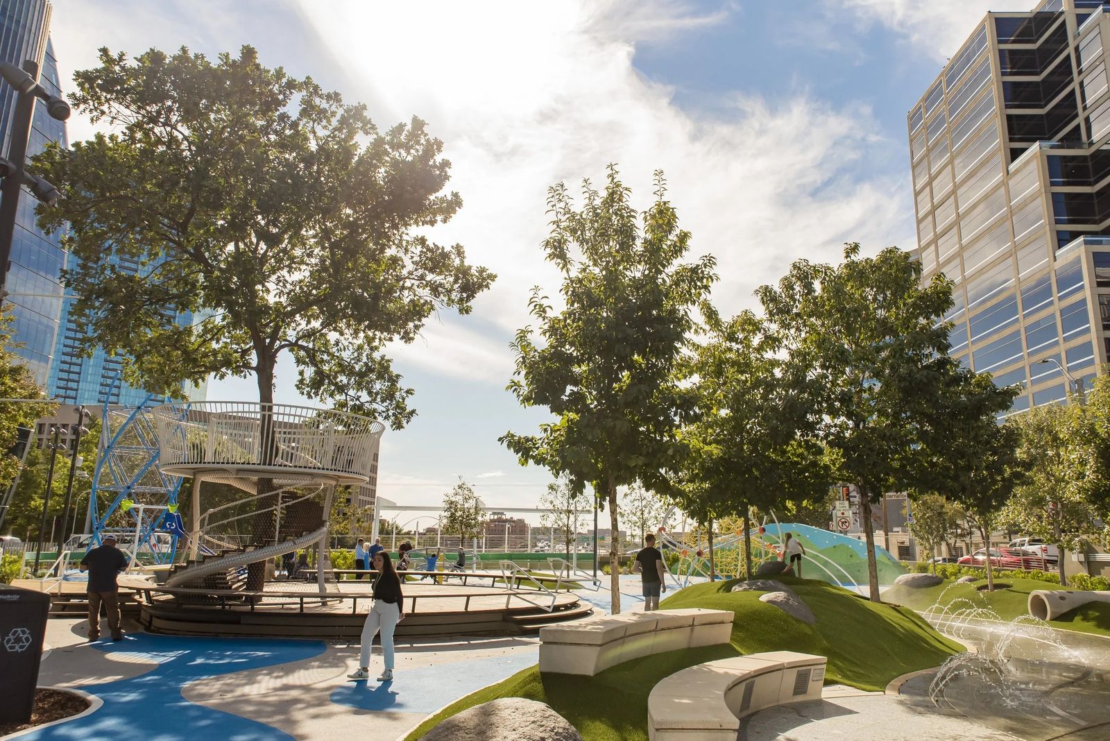 A park with trees, a blue play area and a stage with city buildings rising behind.