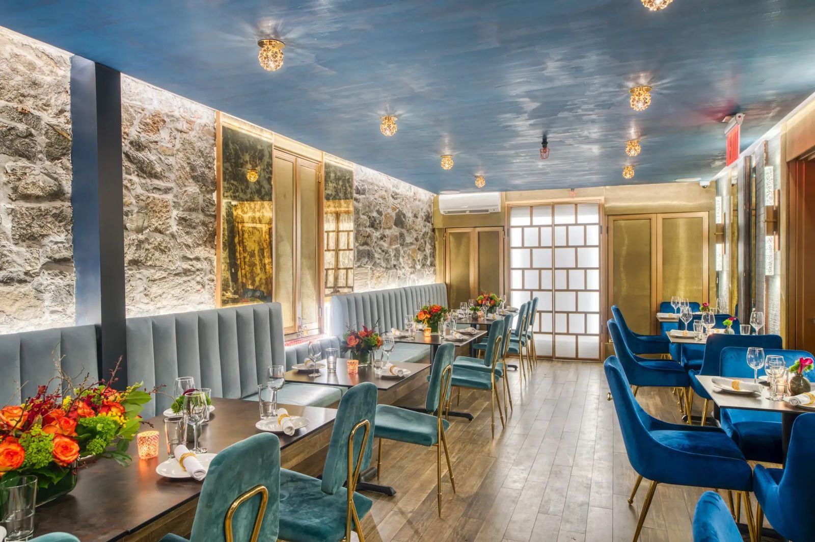 A dining room with a painted sky ceiling, stone wall, blue velvet chairs and tables set with flowers