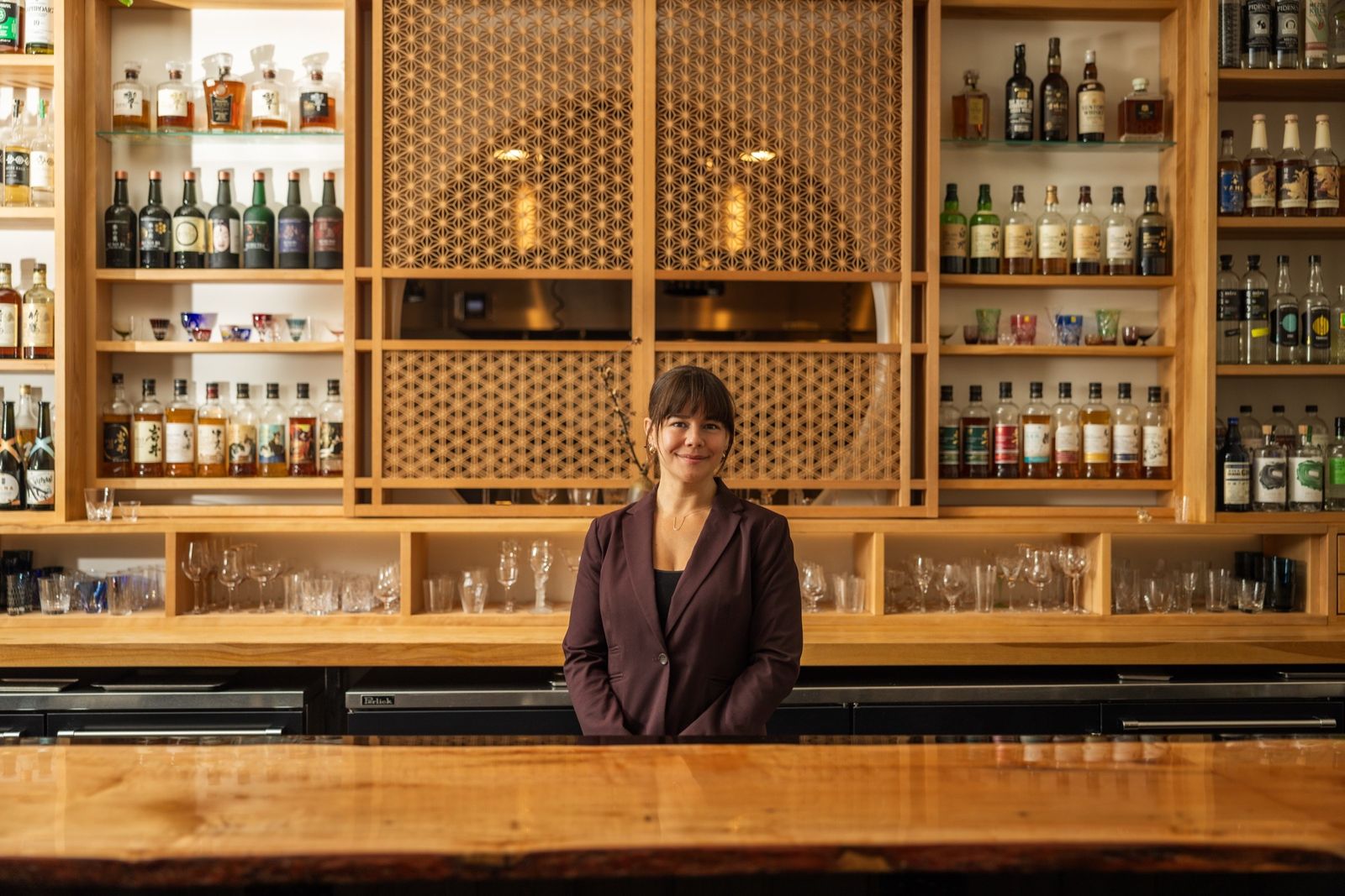 A smiling woman behind a wooden bar in front of a lattice-backed shelf of spirits and glasses.