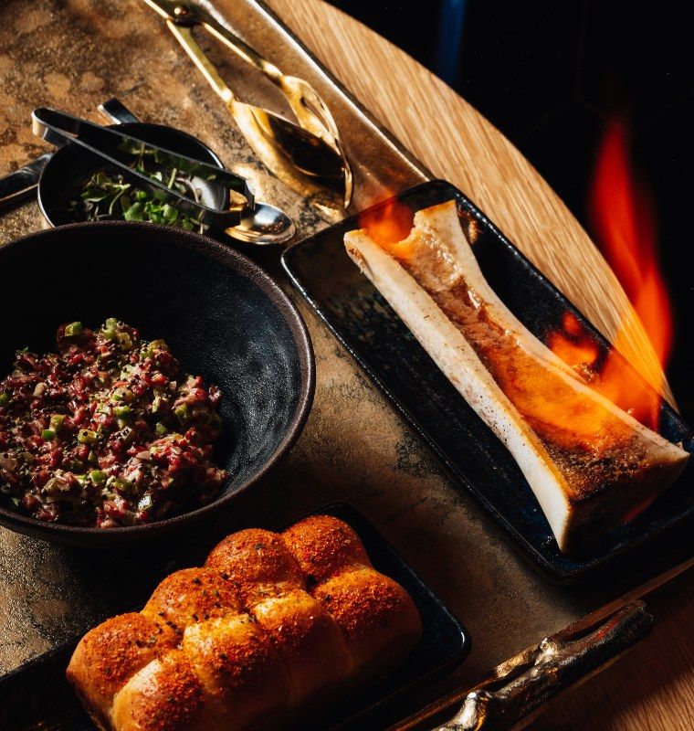 Flaming bone marrow dish with bread rolls and small plates on a wooden table