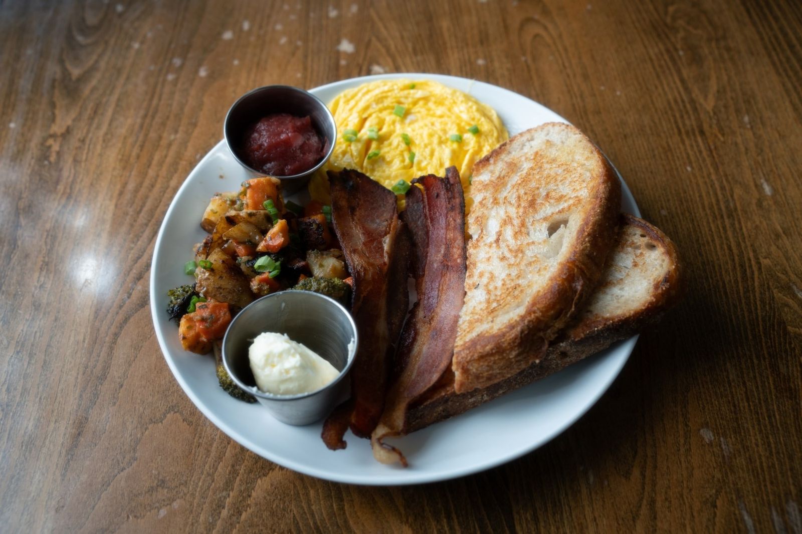 A white plate of scrambled eggs, bacon, roasted potatoes and toast with a small cup of sauce.