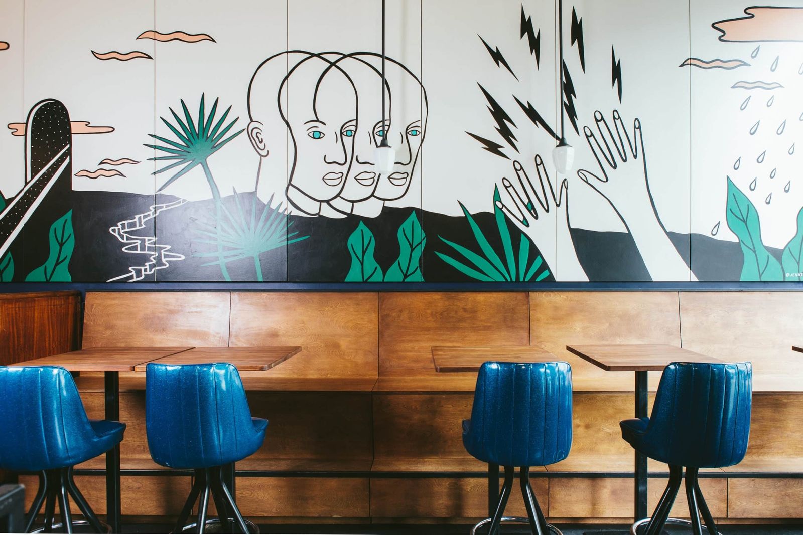 A bar with a painted mural of faces and hands above a wooden bench and bright blue stools.
