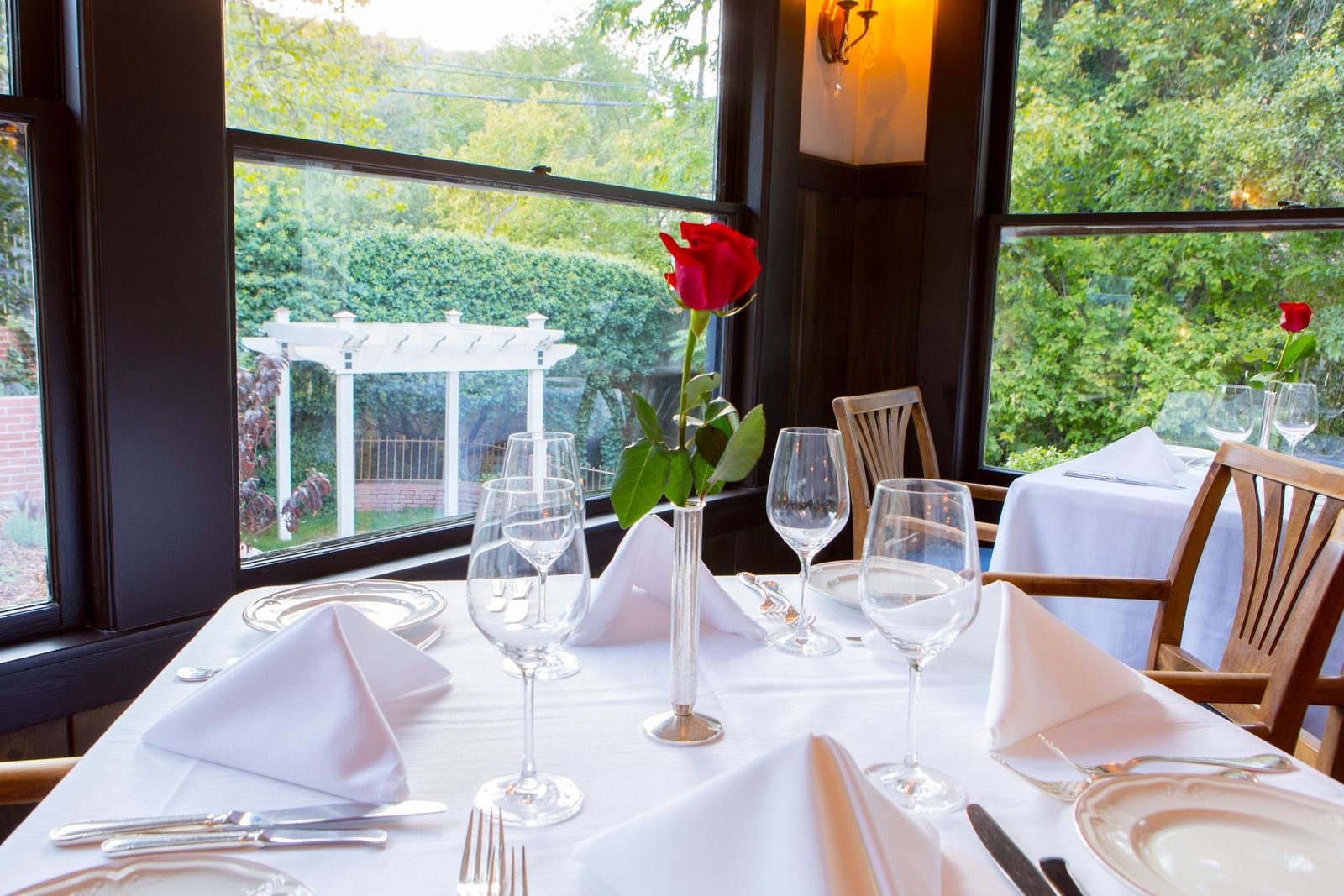 A white-clothed table set with wine glasses and a single red rose beside a window overlooking a garden pergola
