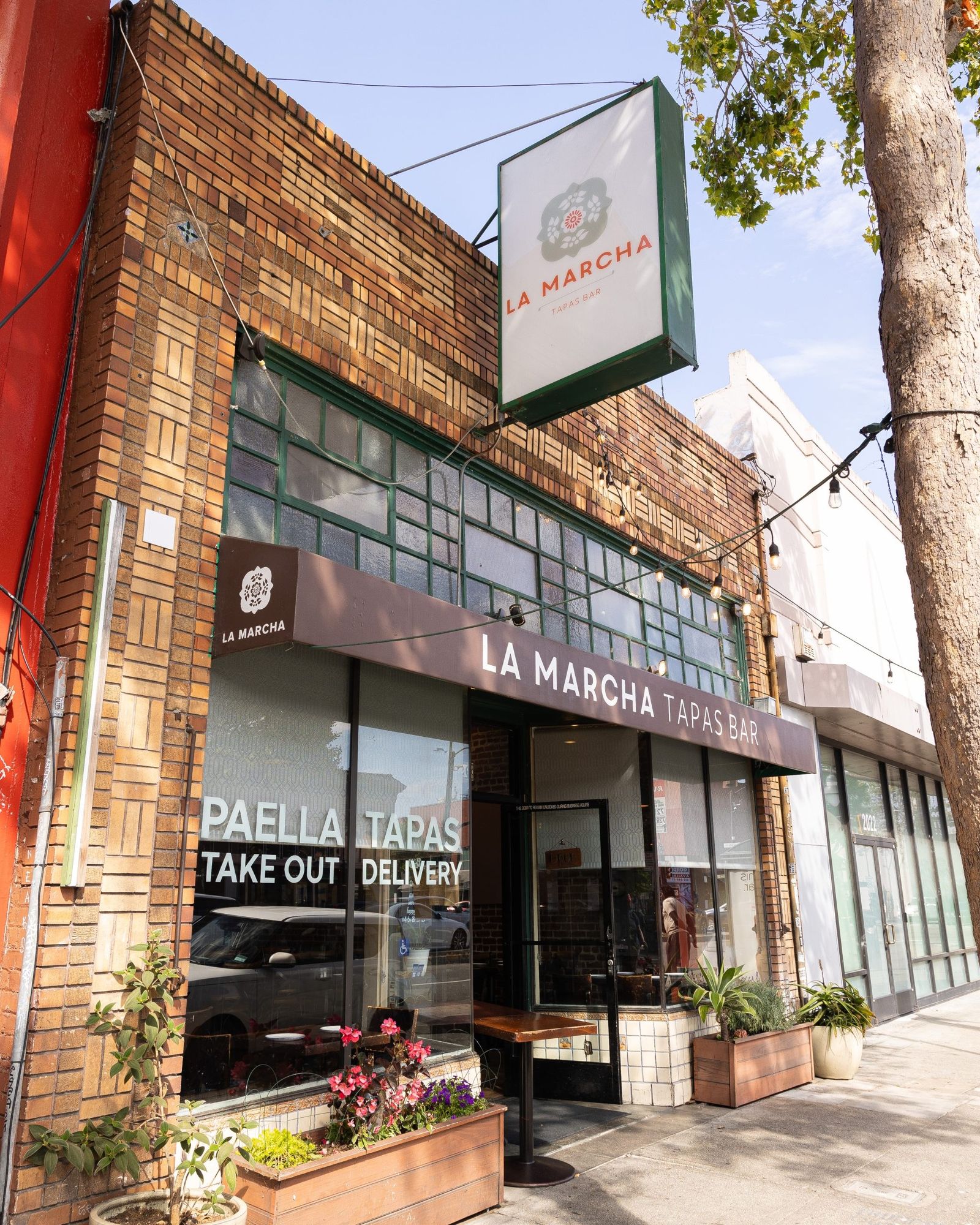 A brick storefront with a green hanging sign reading La Marcha Tapas Bar and a take-out window.