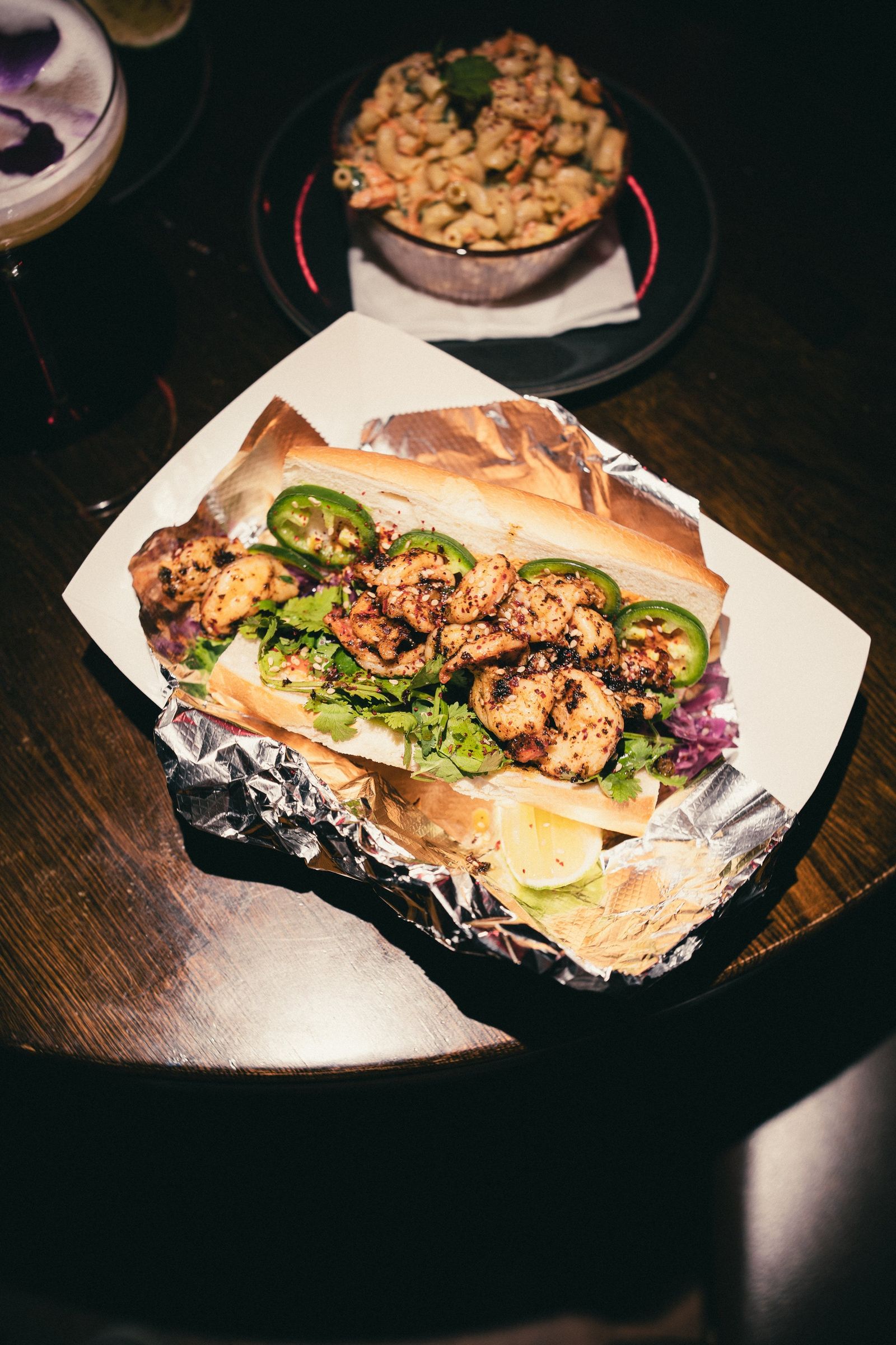 A sandwich with grilled chicken, jalapeno slices and greens in a paper-lined basket on a dark table, with a small dish behind it.