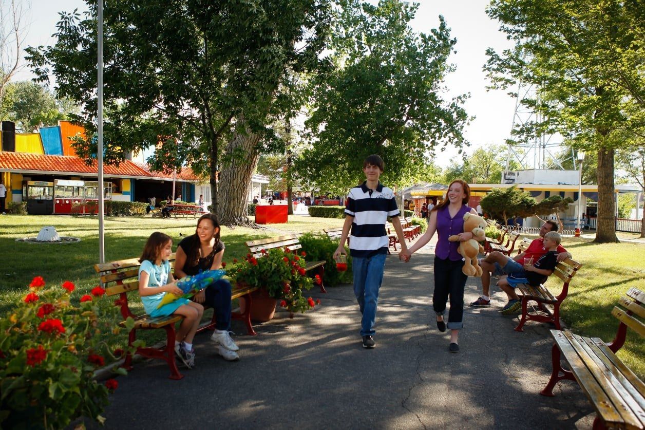 A family walking along a path lined with flower beds and shade trees past colorful amusement rides.