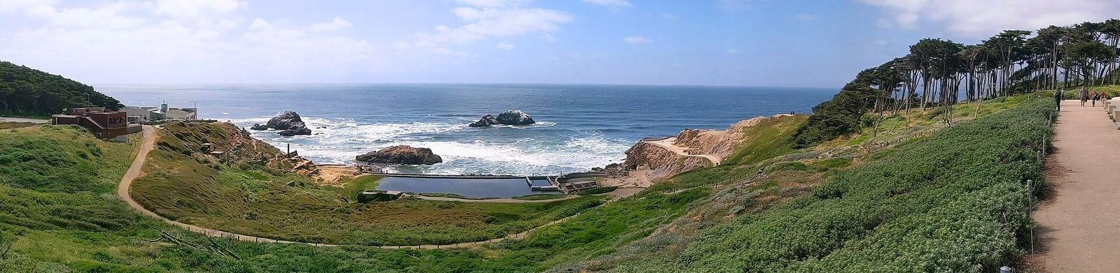 A coastal path winding across a green headland above rocks and surf, with cypress trees on the right.