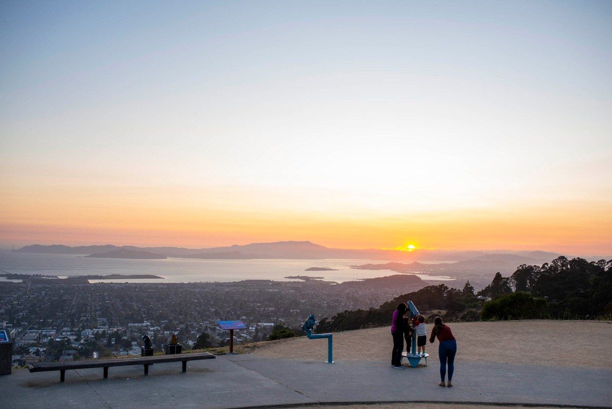 A sunset sky over the bay seen from a hilltop terrace with a few people standing at the edge.