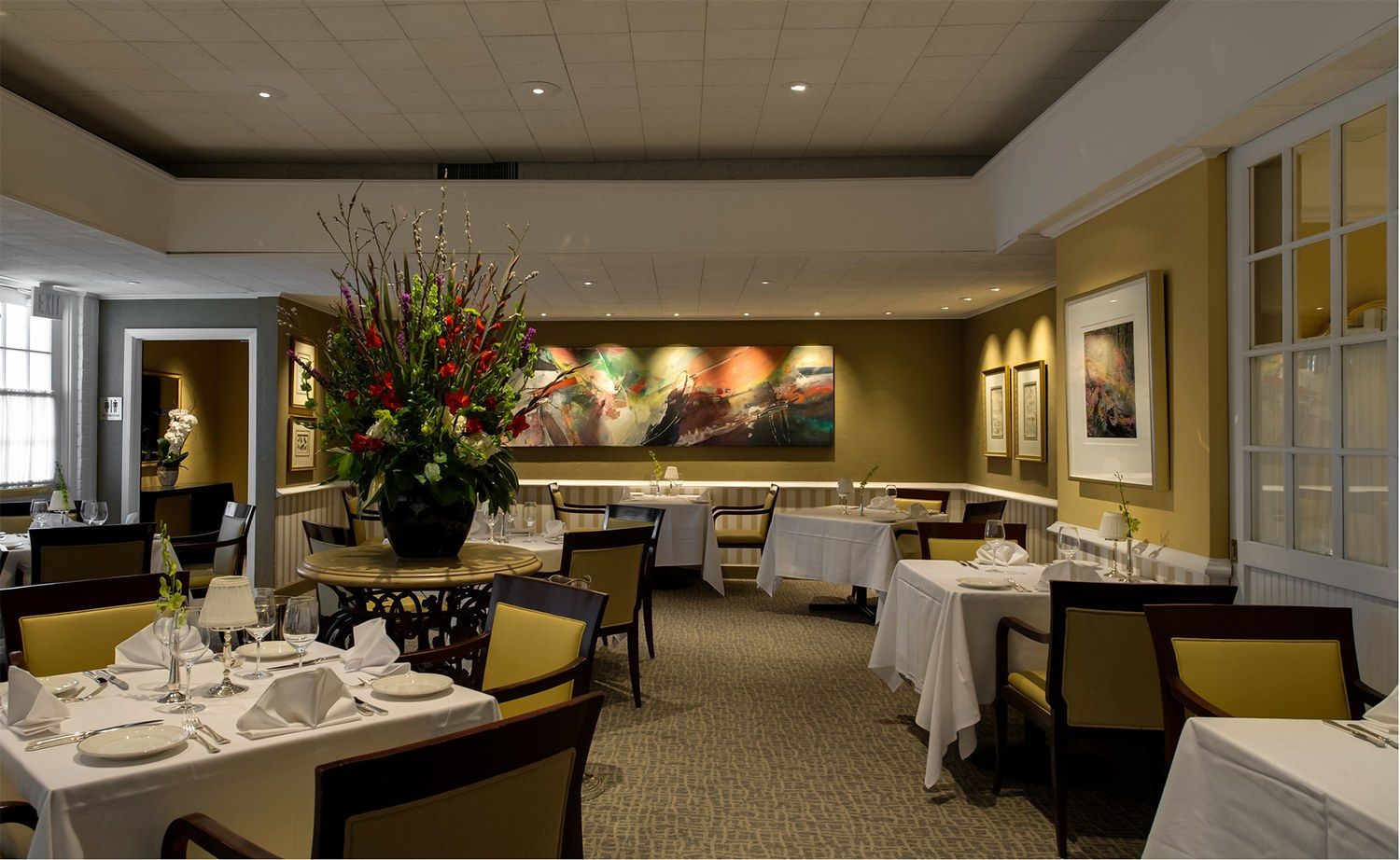 A formal dining room with white-clothed tables, upholstered chairs and a large flower arrangement