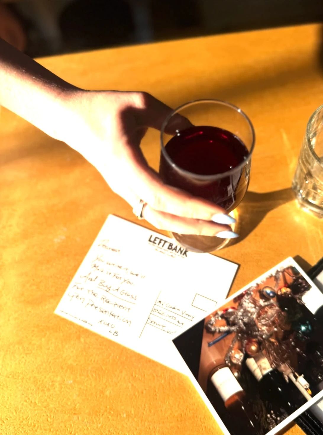 A hand holding a glass of red wine over a postcard on a yellow table, with a printed photo beside it.