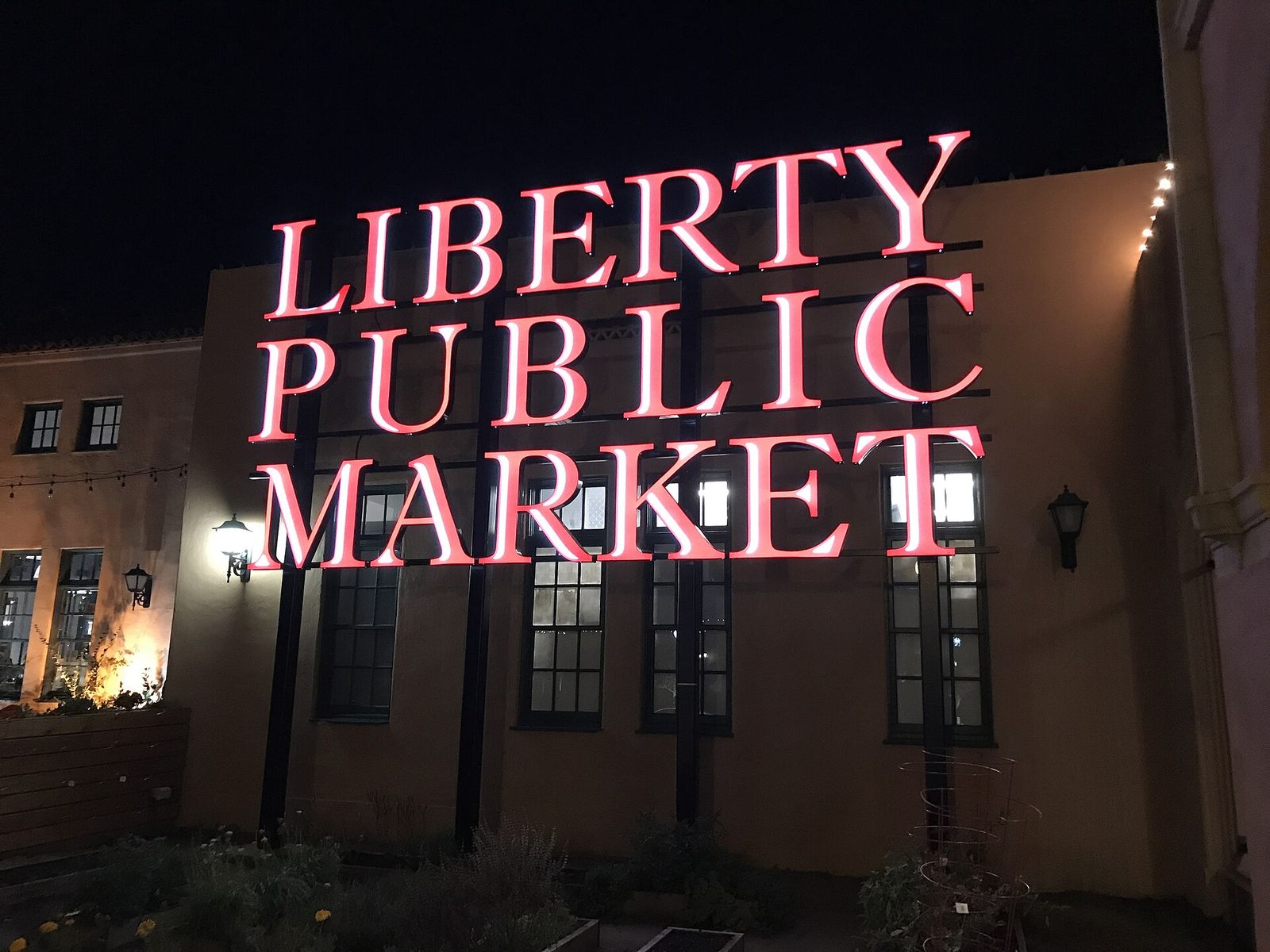 A lit sign reading Liberty Public Market on a stucco building at night.