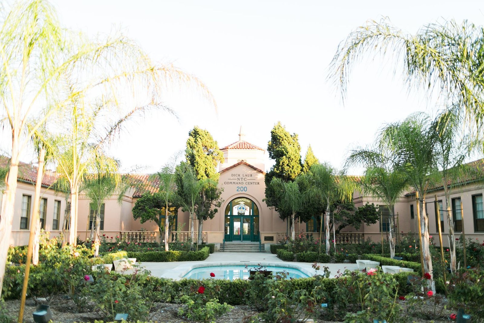 A Spanish-style building with a tower and palm trees behind a fountain and rose garden.