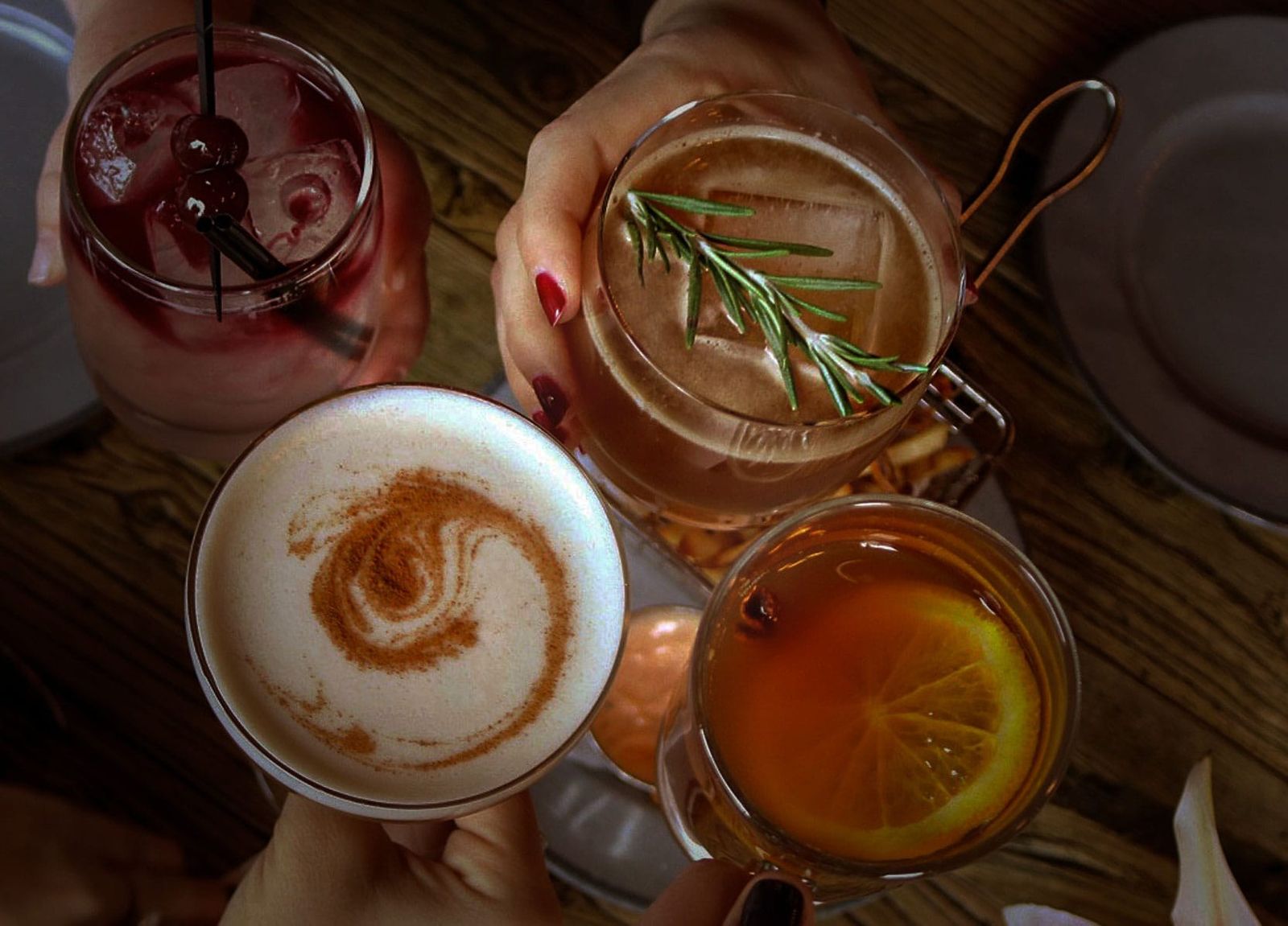 Hands around several cocktails on a dark wooden table, one with a rosemary sprig and one topped with foam.