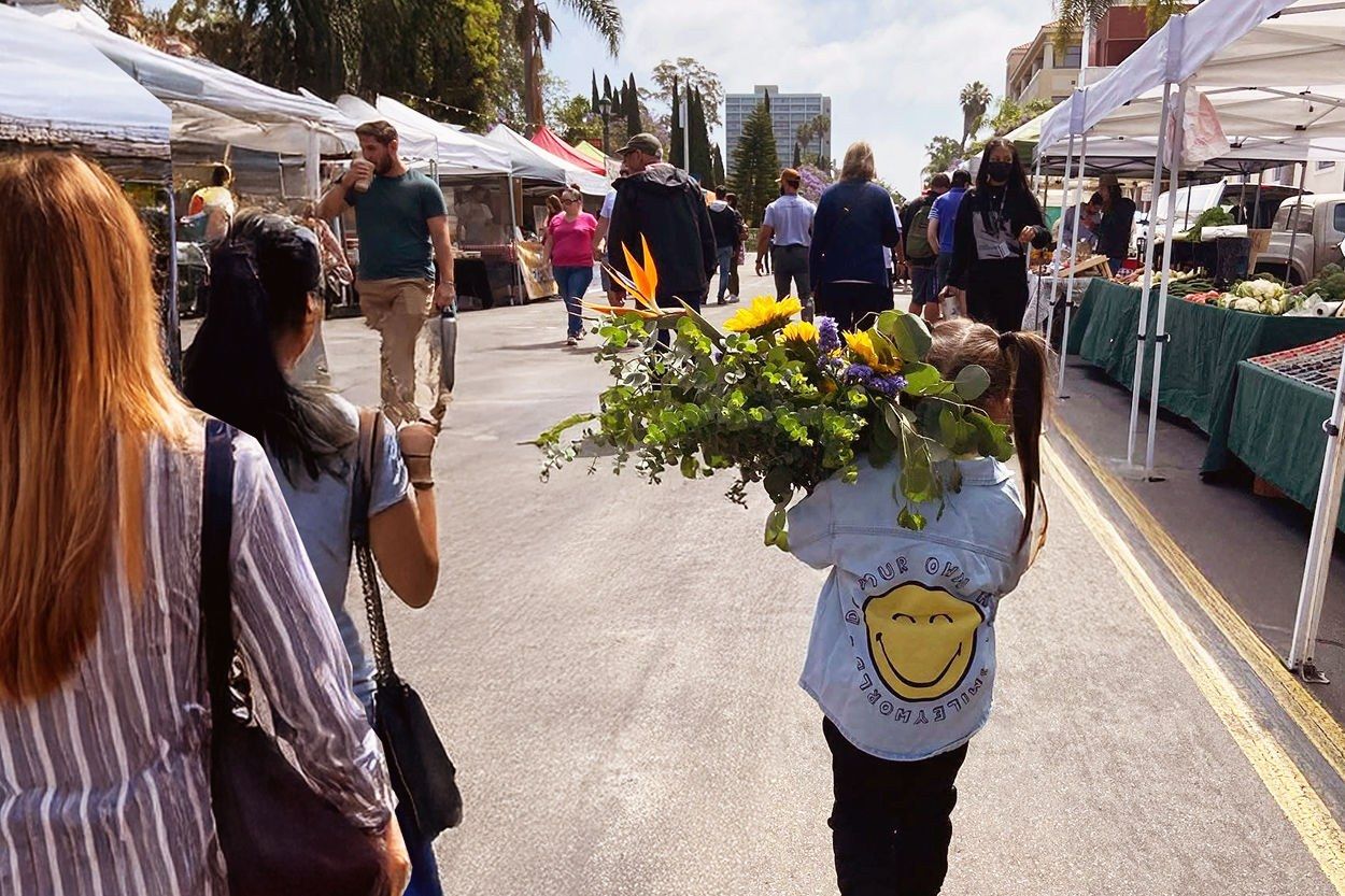 A farmers market street lined with white tents and shoppers, with one carrying yellow flowers.