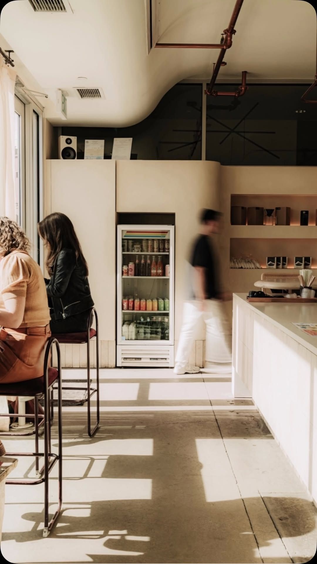 A sunlit cafe counter with a few stools and customers, a drinks cooler and shelves at the back.