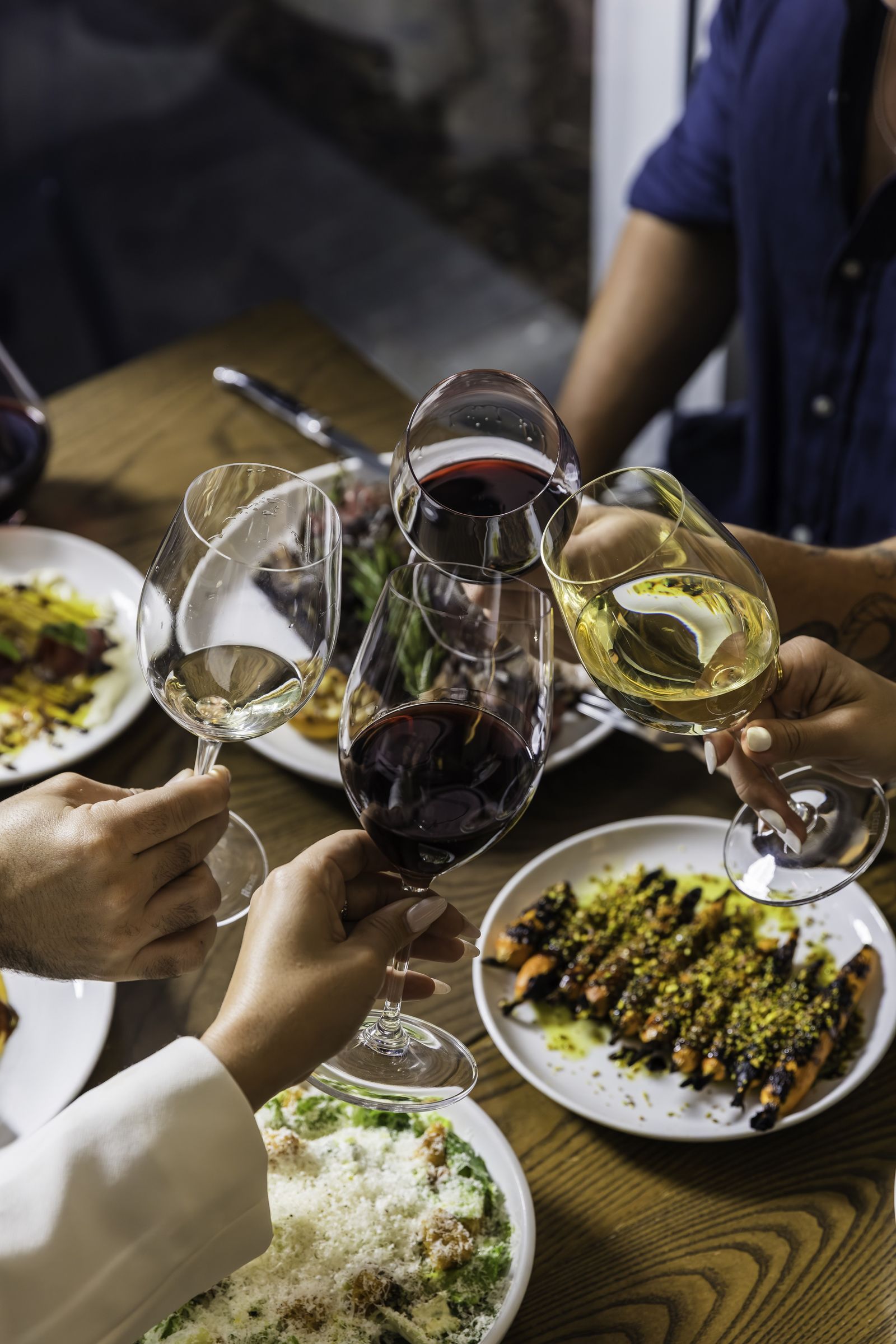 Hands clinking wine glasses over plates of food at a wood table
