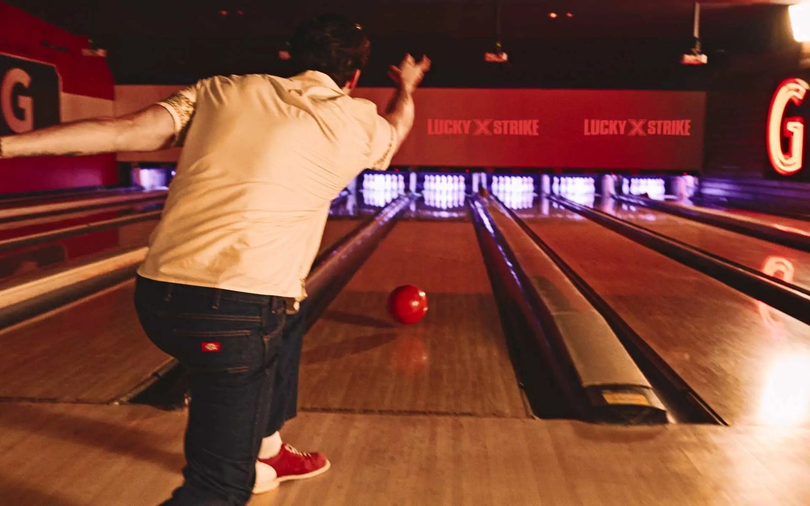A bowler in a tan shirt releasing a ball down a lit bowling lane toward red signage at the far end.
