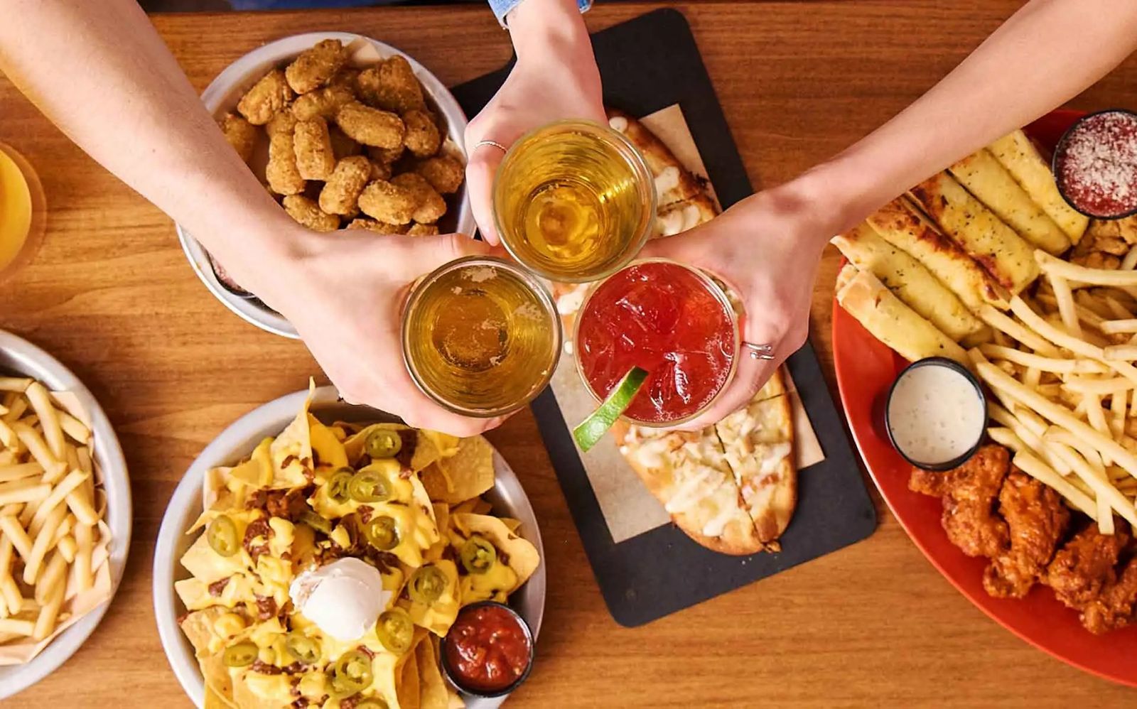 Hands reaching for plates of fries, wings and nachos and glasses of drinks on a wooden table.