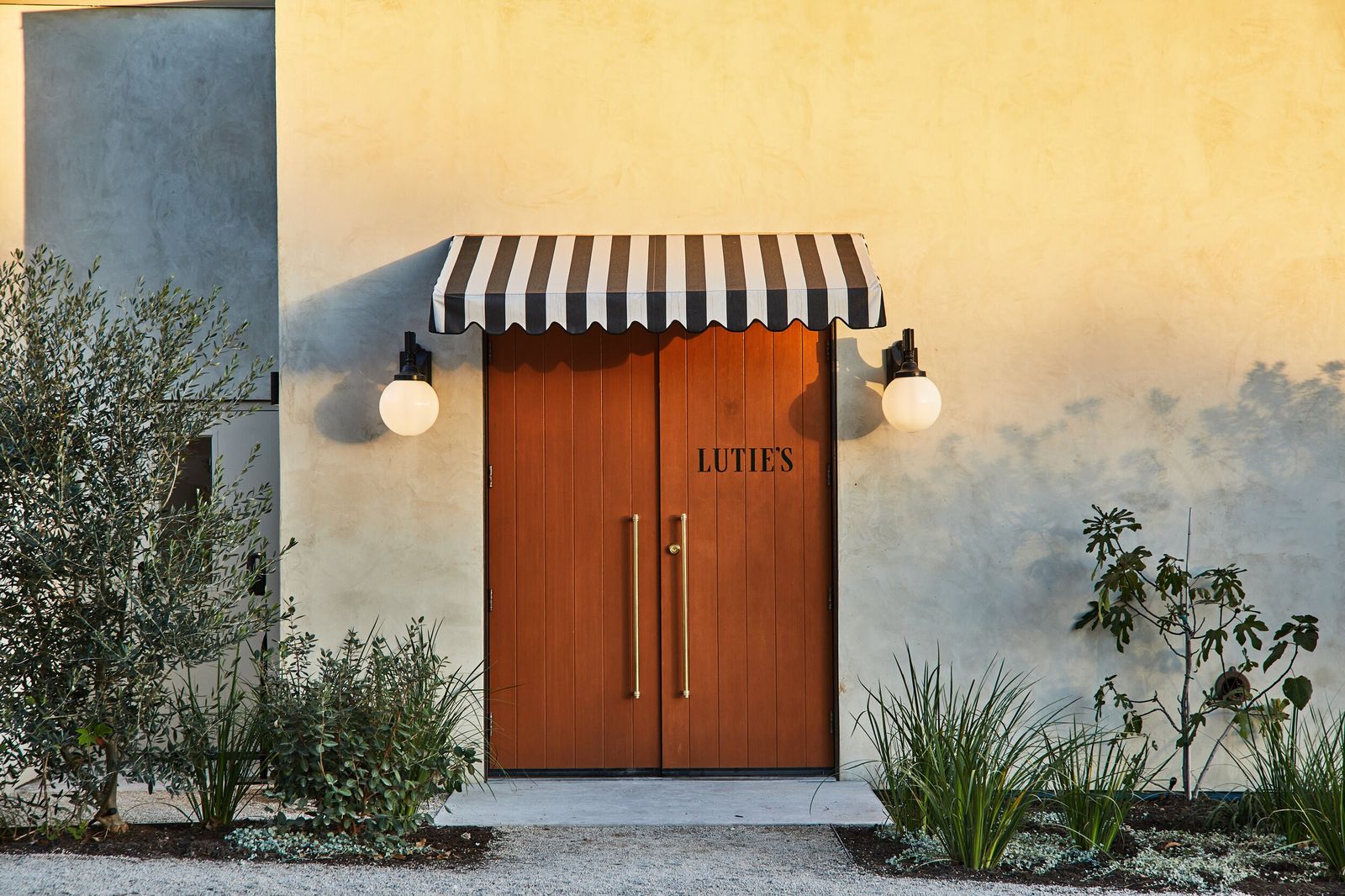 Wooden double doors beneath a black-and-white striped awning, with globe sconces and plants at the entrance.