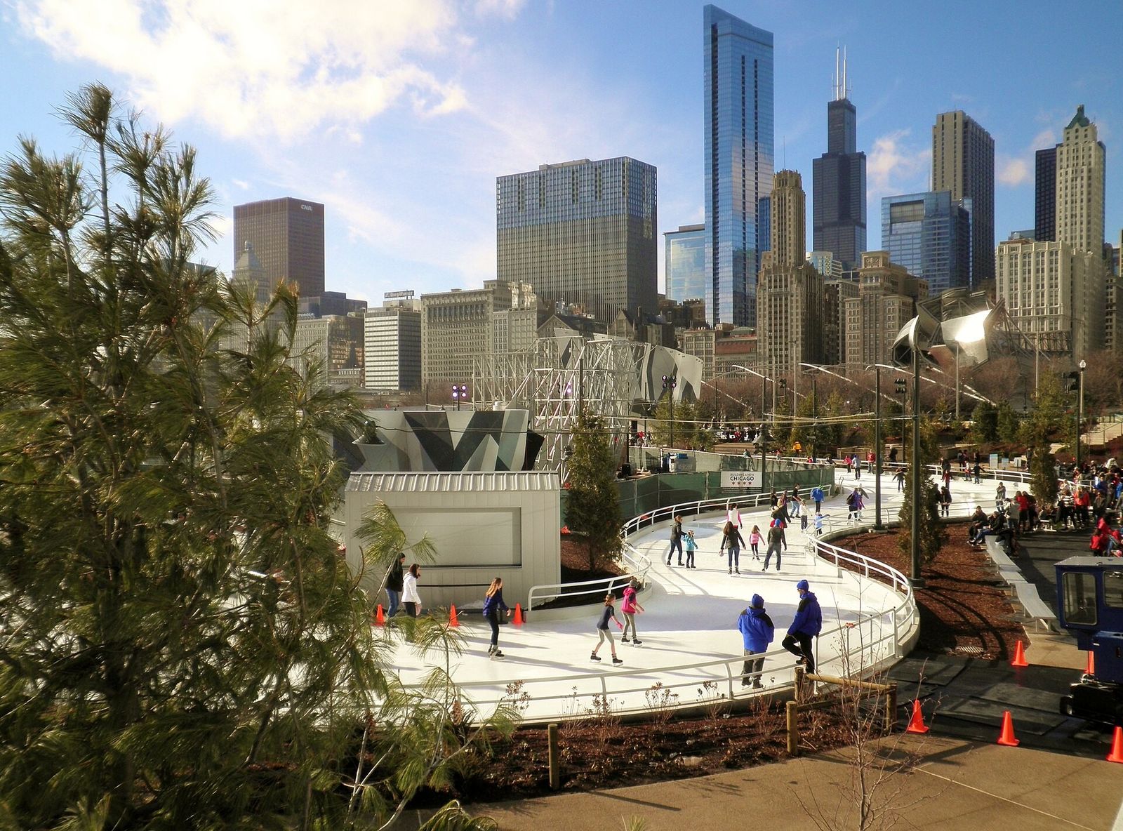A curving ice skating ribbon with skaters and a view of downtown towers beyond the trees.