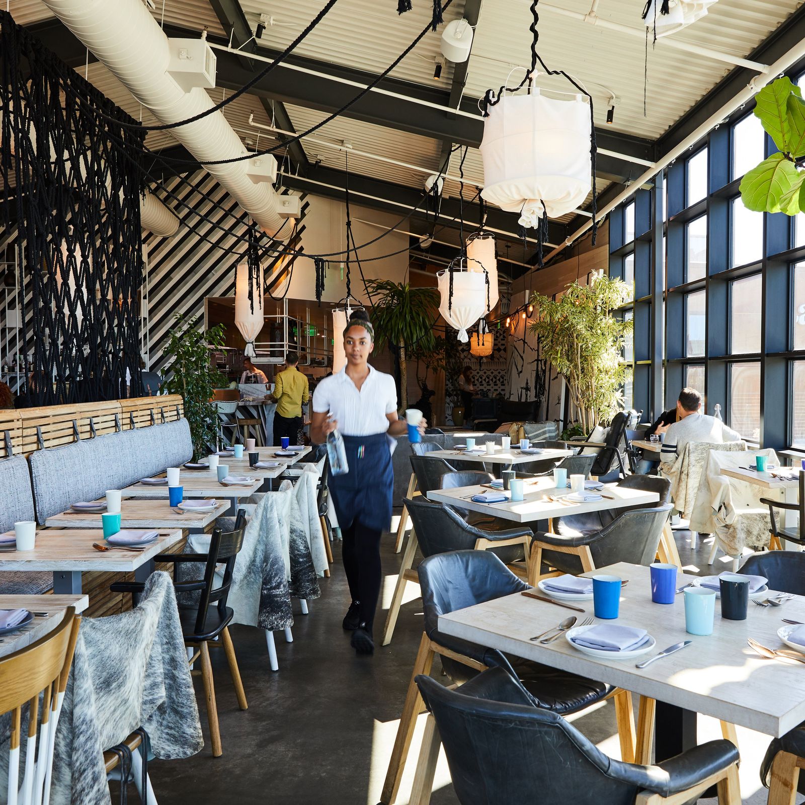 Bright dining room with blue-gray chairs, white tables and large windows, a server walking between tables