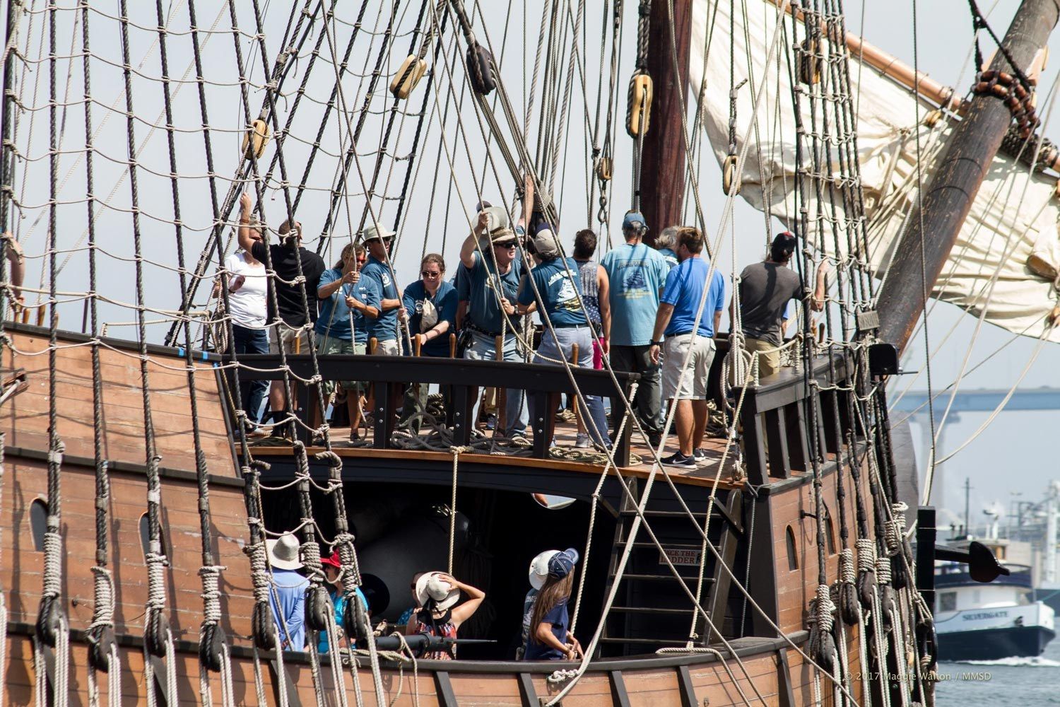A tall ship's rigging and deck crowded with visitors, with the harbor behind.