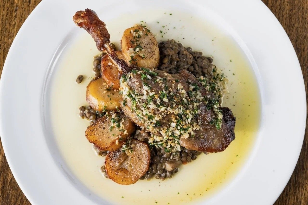 A plate of braised duck leg with lentils and a green sauce on a white plate.