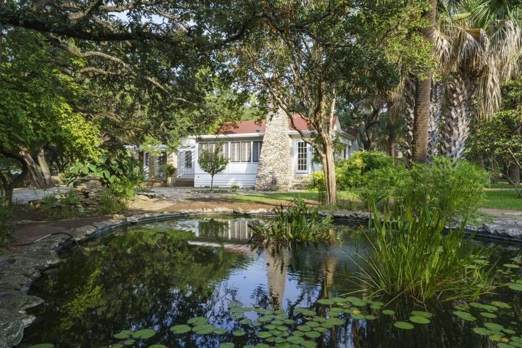 A pond with lily pads in front of a stone cottage among tall trees.