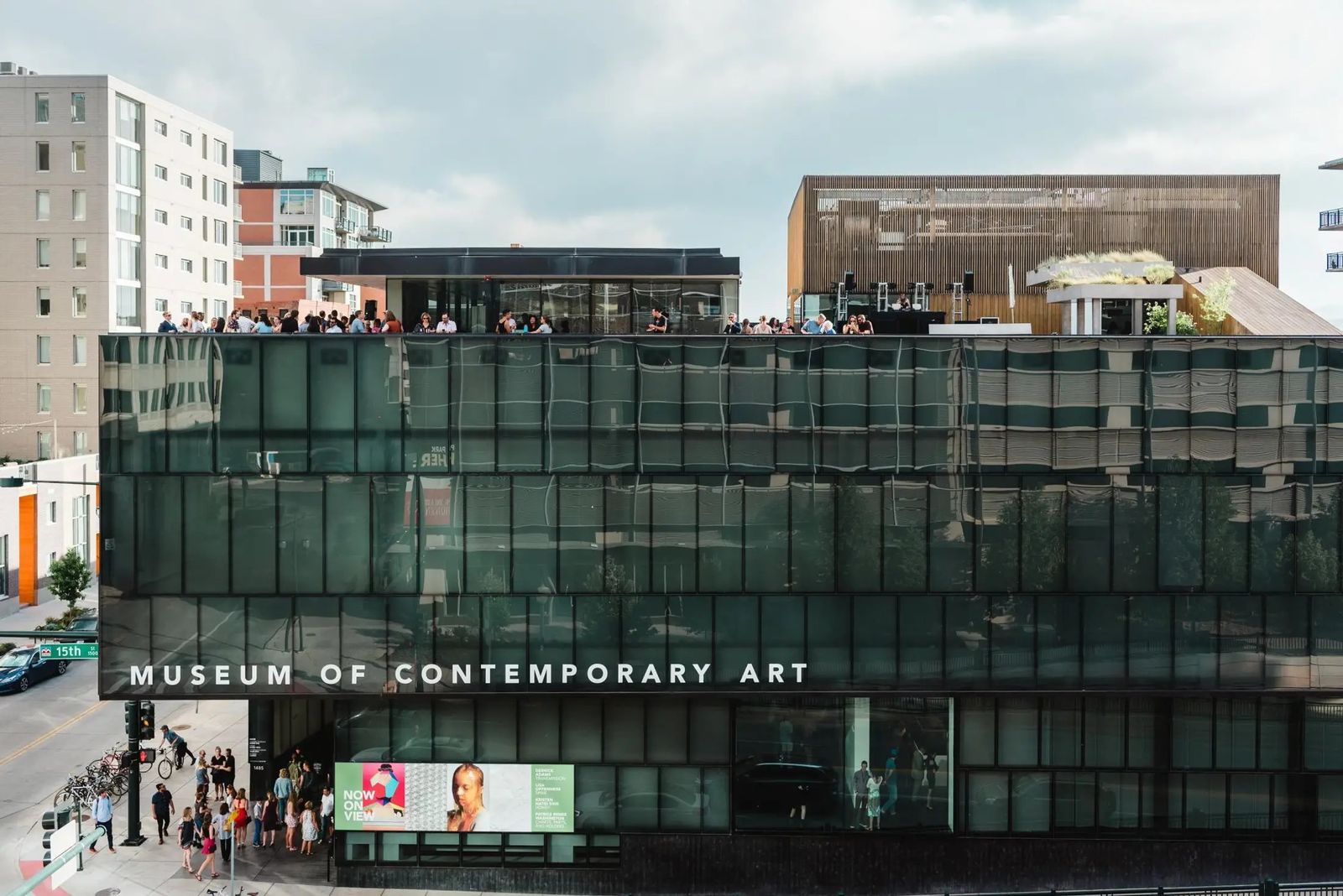 The museum's dark glass and metal building with a rooftop crowd and a wall sign reading Museum of Contemporary Art.
