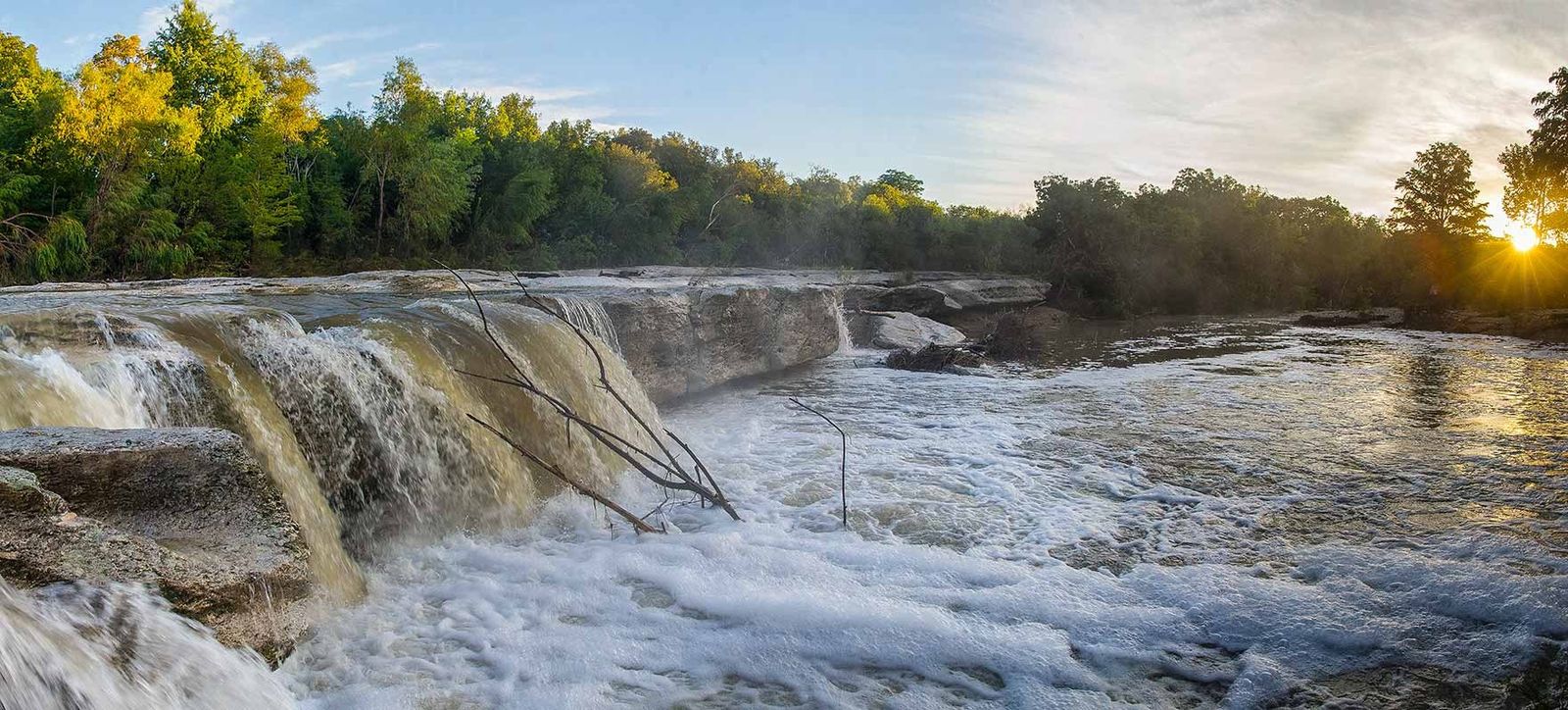 A low waterfall running over limestone ledges at sunset with trees along the bank.