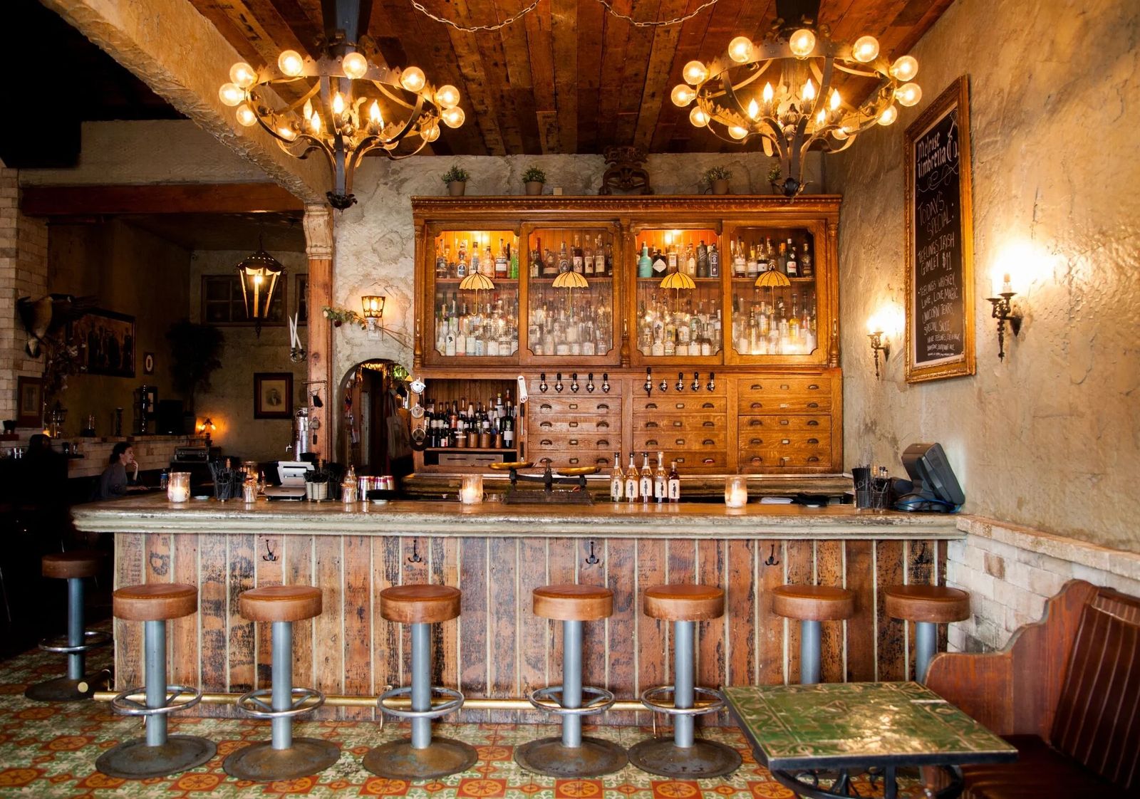 A bar with a wooden counter, stools, shelves of bottles and brass chandeliers over a tiled floor.