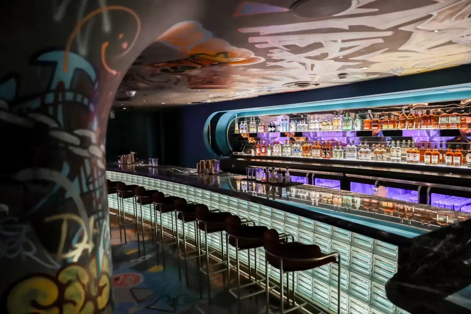 A long curving bar with a lit countertop, purple stools and graffiti-covered walls in a dark room.