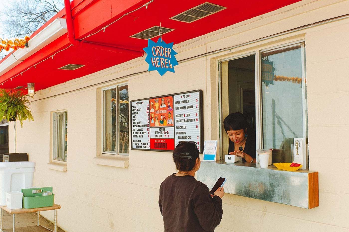 A red-trimmed order window on a cream building with a person ordering at the window.