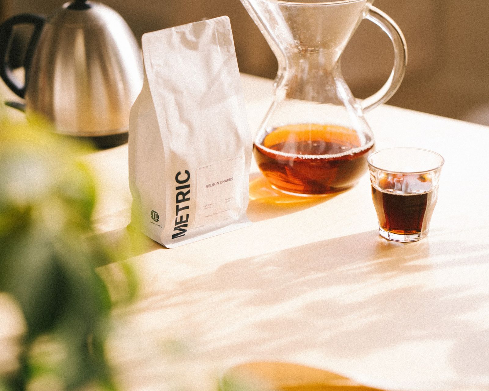 A white Metric coffee bag beside a glass pour-over server and a small cup of coffee on a sunlit table.