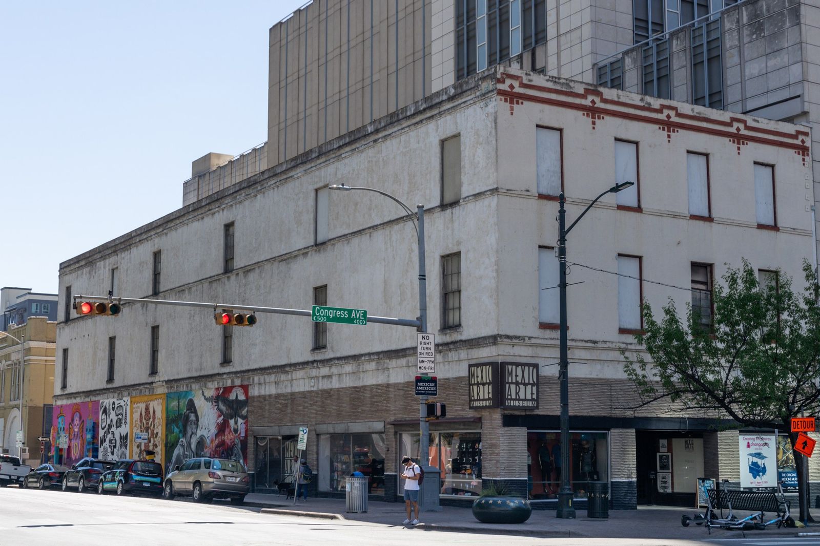 A multi-story stone building at a street corner with a mural along its ground floor and traffic lights.