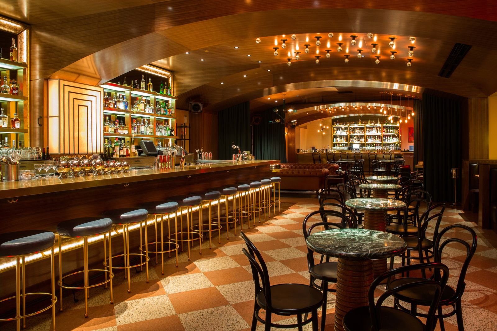 A long bar with stools beside a checkerboard-floored room with round tables and an arched ceiling, lit in warm amber.