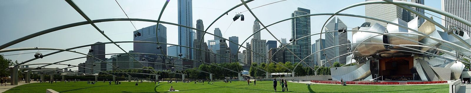 A wide panorama of the Pritzker Pavilion's trellis over the lawn with skyscrapers behind.